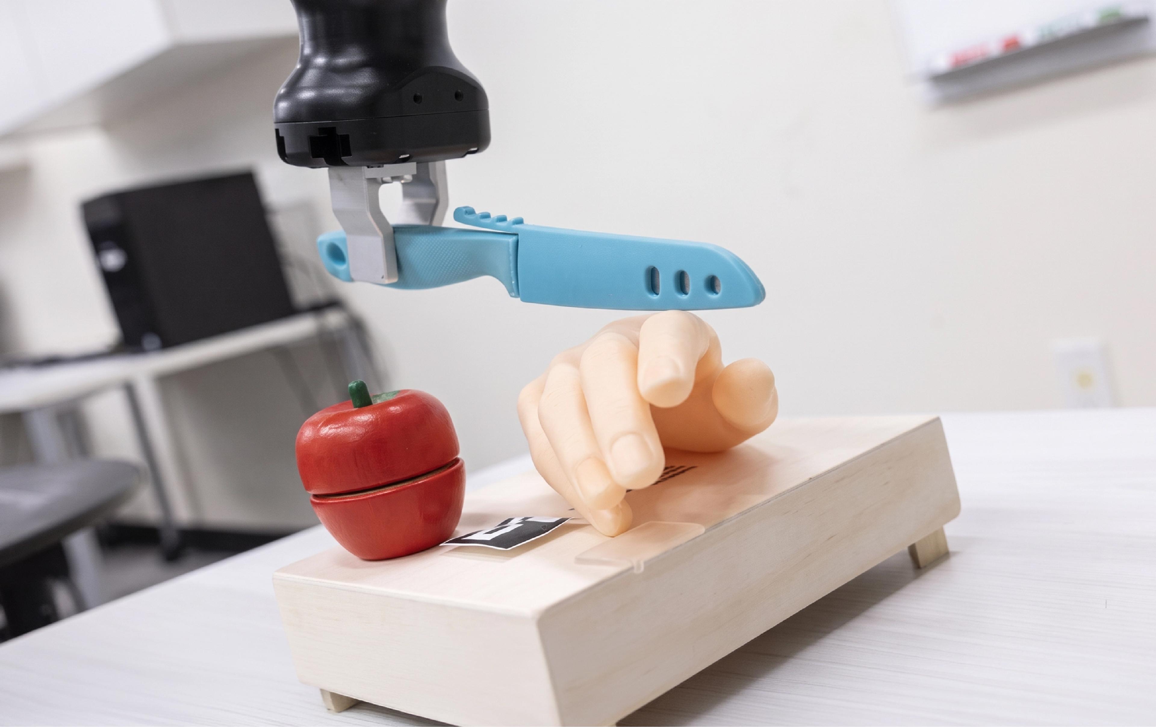 A robotic arm holds a plastic knife above a prosthetic hand and an apple in a lab setting.