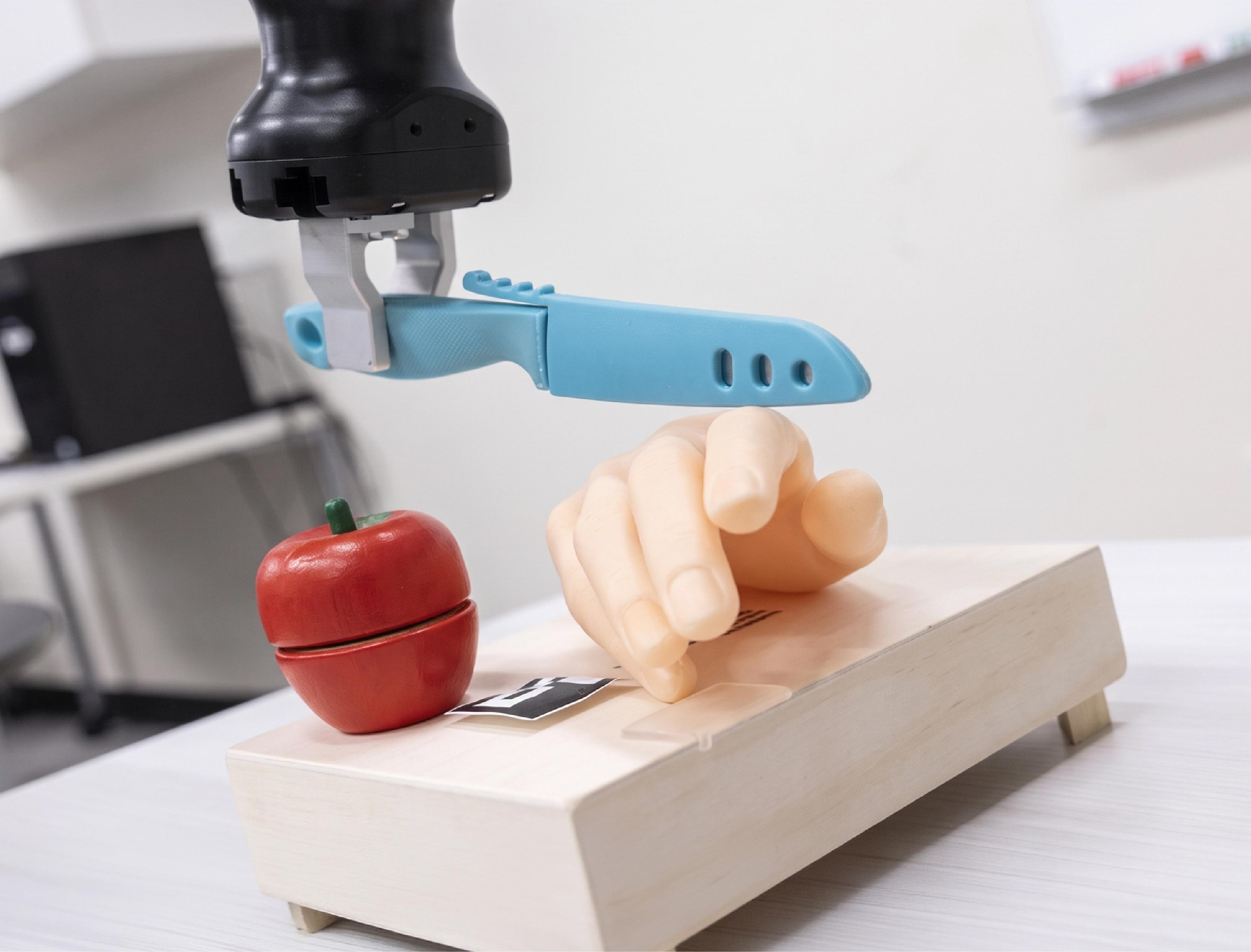 A robotic arm holds a plastic knife above a prosthetic hand and an apple in a lab setting.