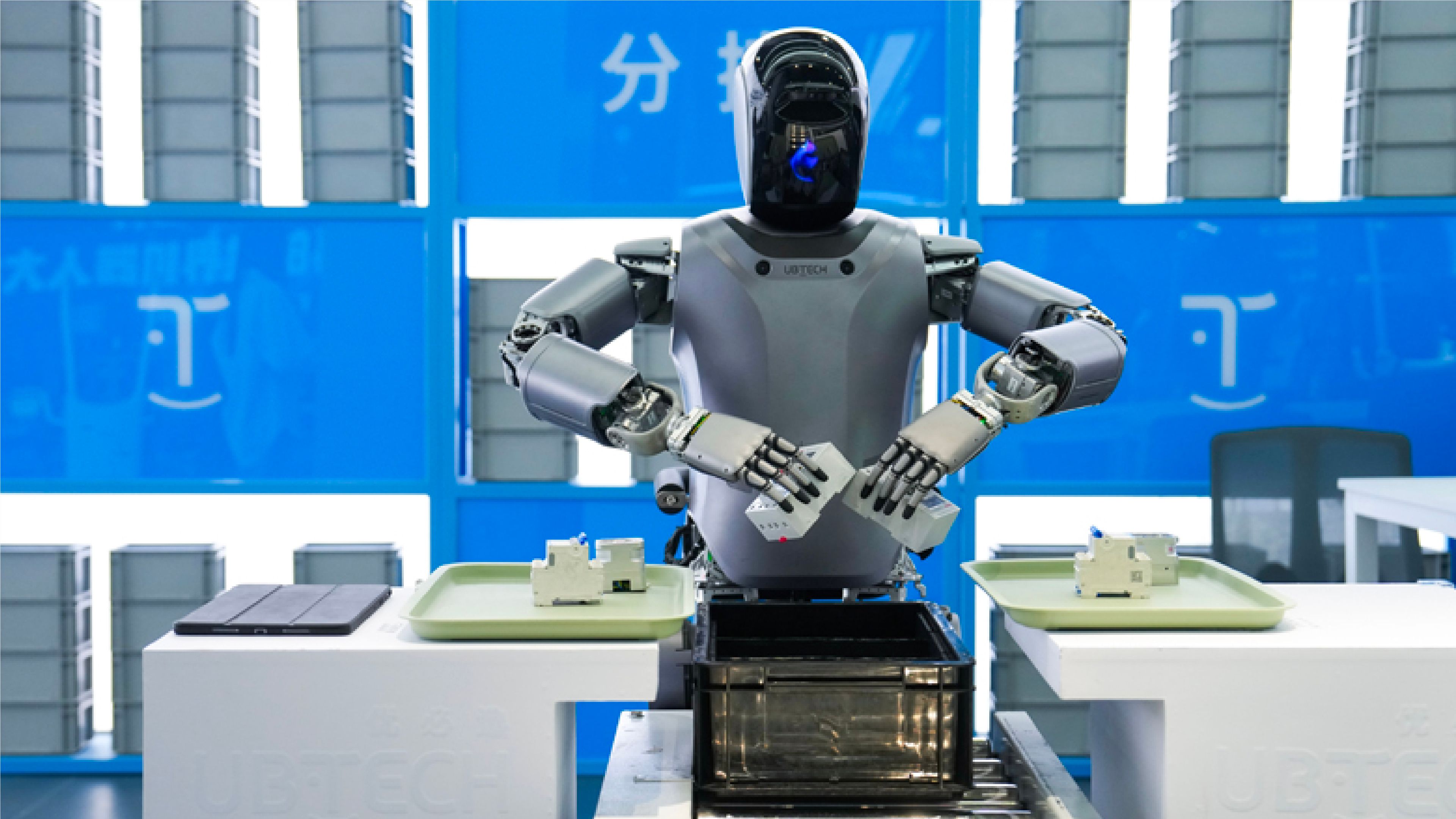 A UBTECH humanoid robot with a black visor-style head and gray torso stands at an industrial workstation, reaching toward small electronic components on trays as it sorts items into a black bin on a conveyor, with stacked gray containers and blue signage in the background.