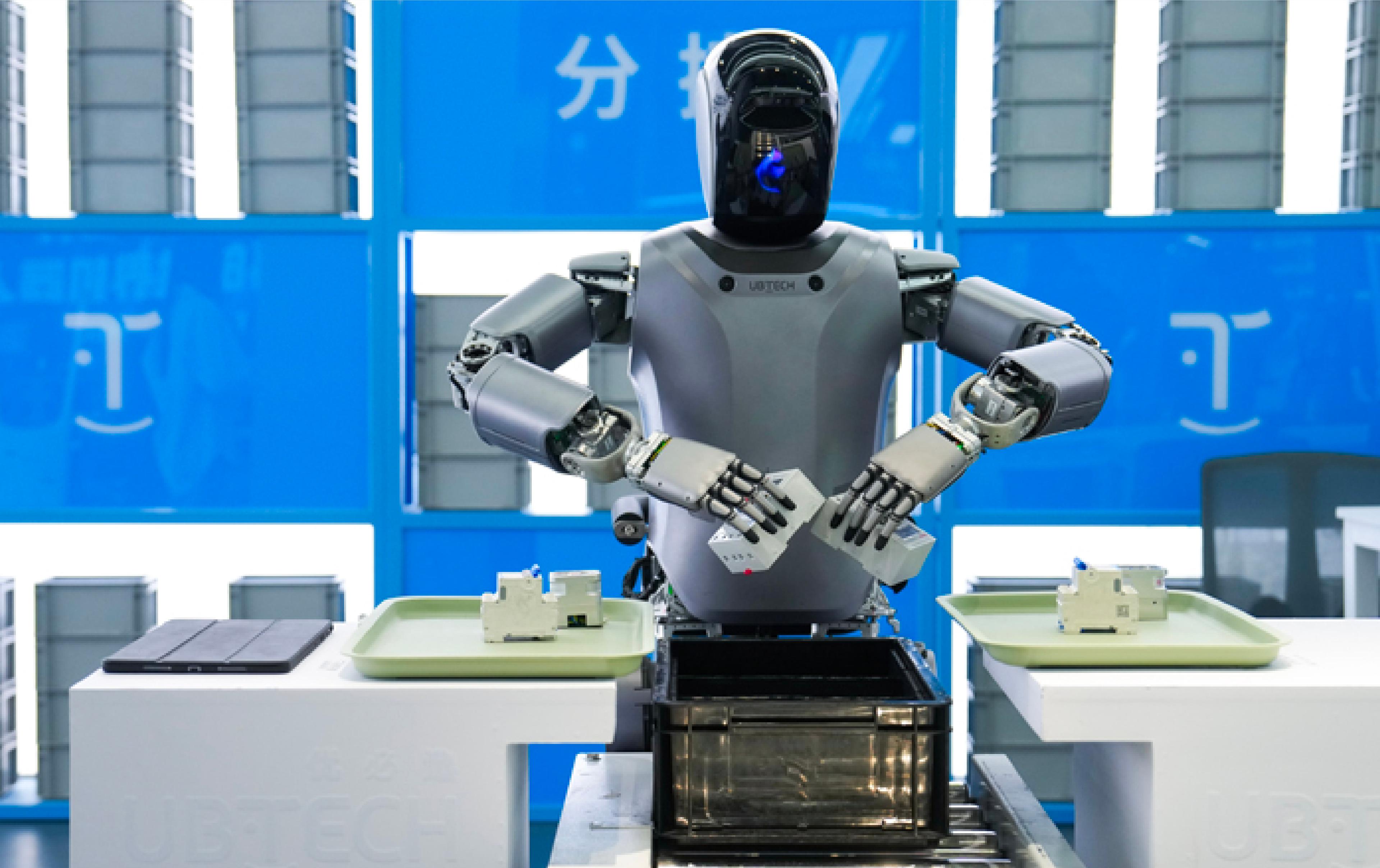 A UBTECH humanoid robot with a black visor-style head and gray torso stands at an industrial workstation, reaching toward small electronic components on trays as it sorts items into a black bin on a conveyor, with stacked gray containers and blue signage in the background.