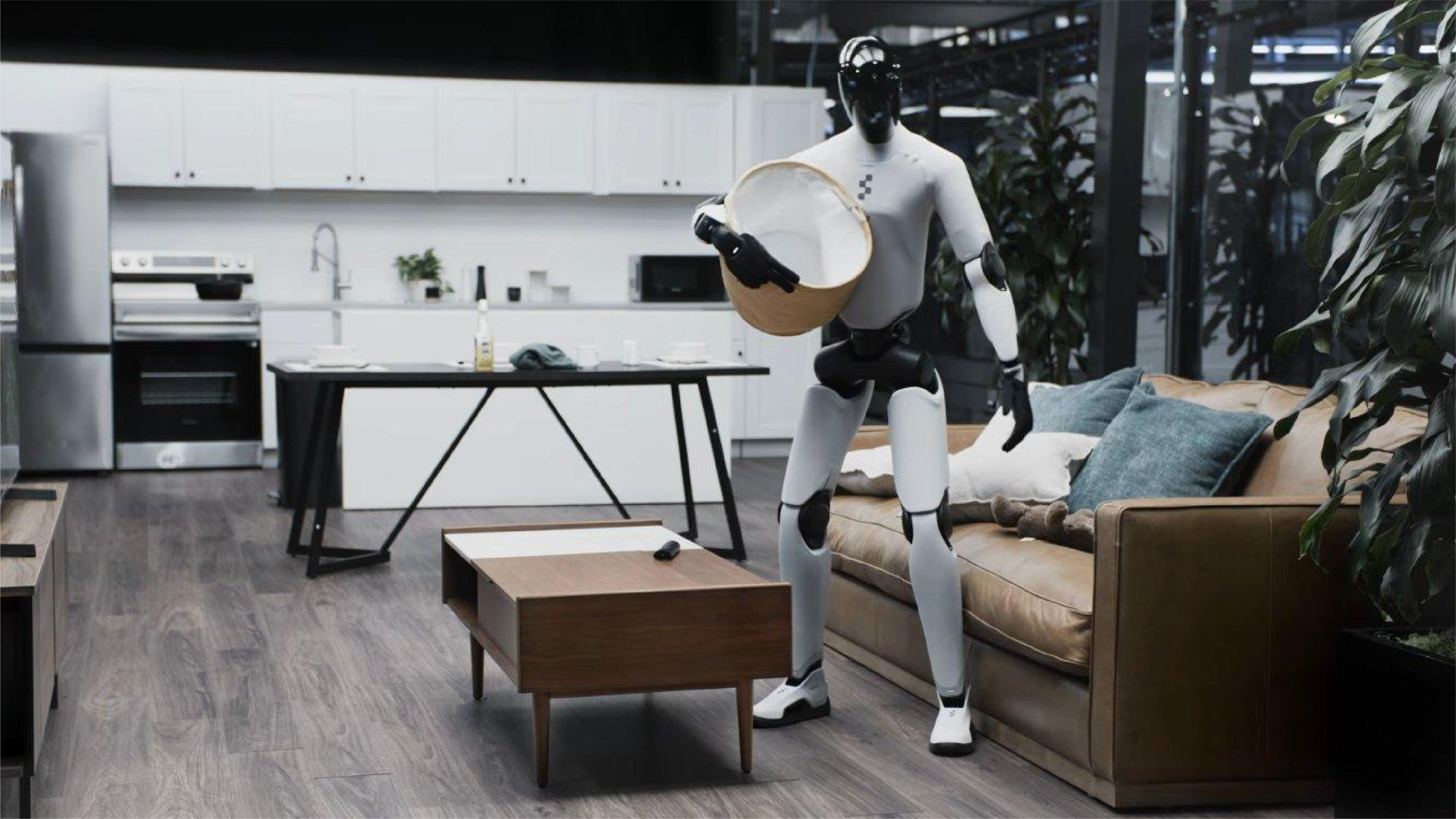 A humanoid robot carries a laundry basket in a modern home setting, standing between a living room sofa and an open kitchen.