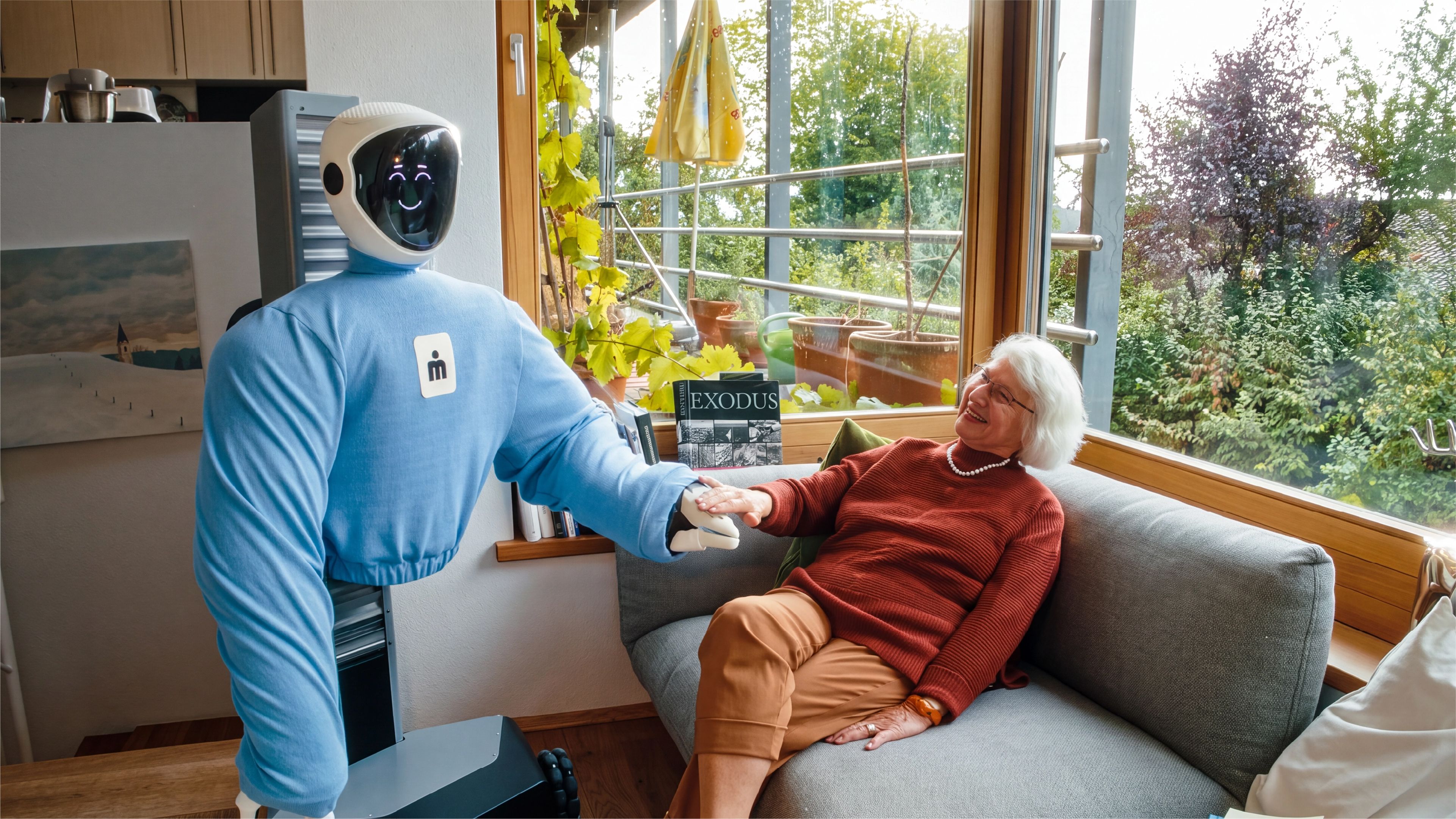 Plim Robotics' wheeled humanoid robot holds hands with an older woman seated on a sofa in a living room.
