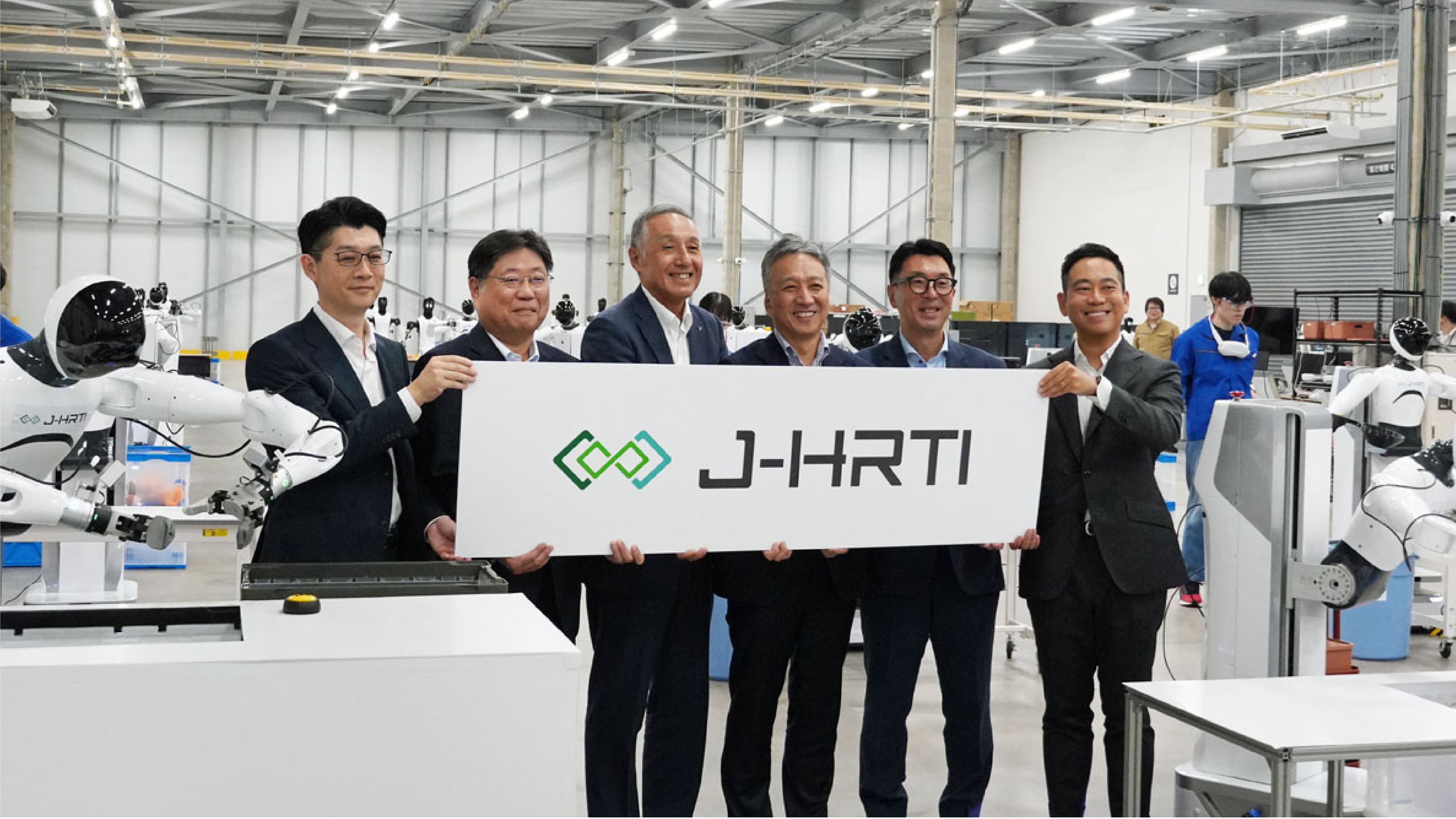 Six executives from J-HRTI's founding companies hold a J-HRTI branded sign at the launch of the Kanto Data Factory in Narashino, Chiba, flanked by white humanoid robots on the factory floor.