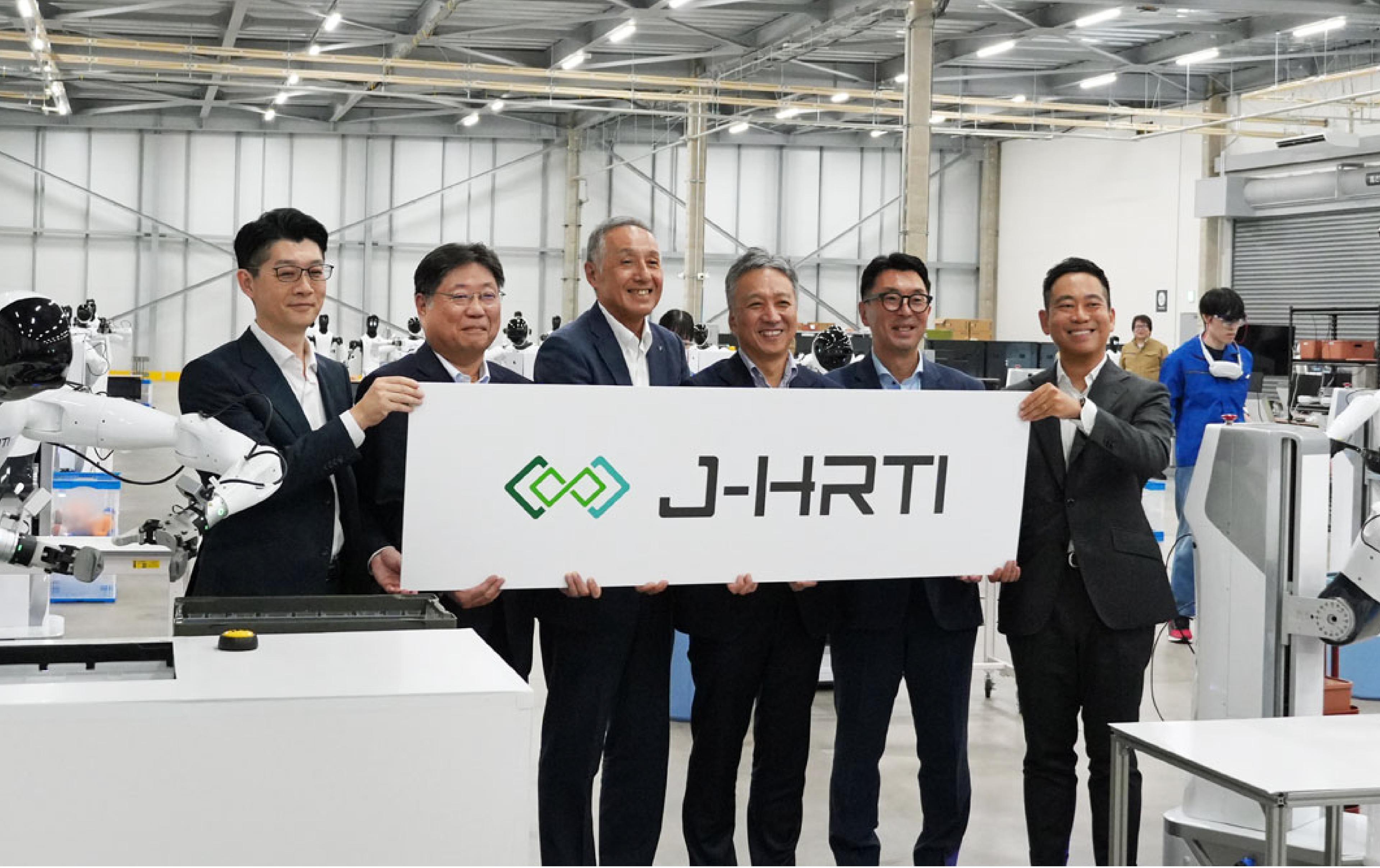 Six executives from J-HRTI's founding companies hold a J-HRTI branded sign at the launch of the Kanto Data Factory in Narashino, Chiba, flanked by white humanoid robots on the factory floor.