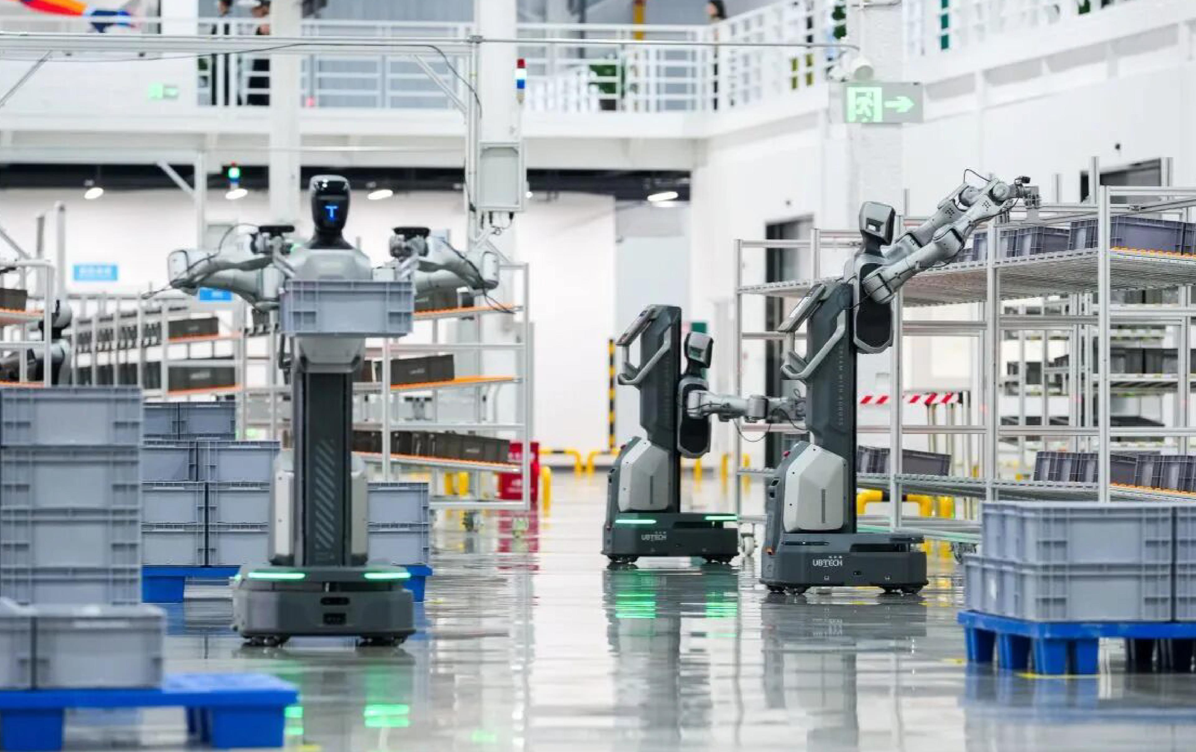 UBTECH humanoid robots with wheeled bases and articulated arms operate among storage racks and plastic bins in an industrial logistics facility.