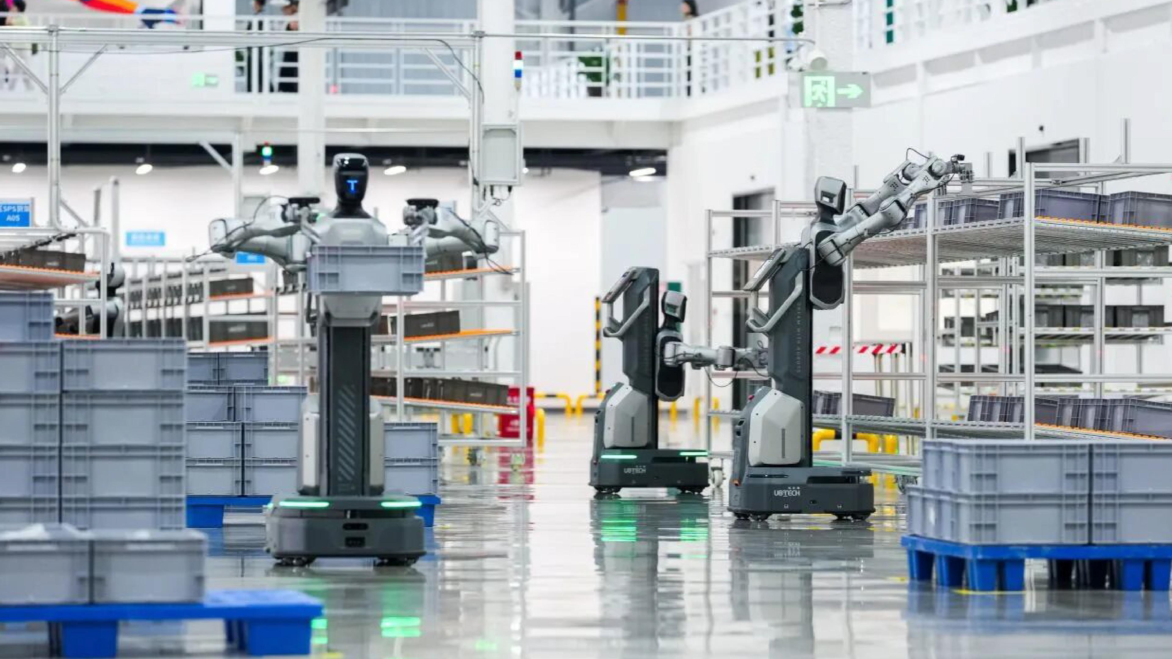 UBTECH humanoid robots with wheeled bases and articulated arms operate among storage racks and plastic bins in an industrial logistics facility.