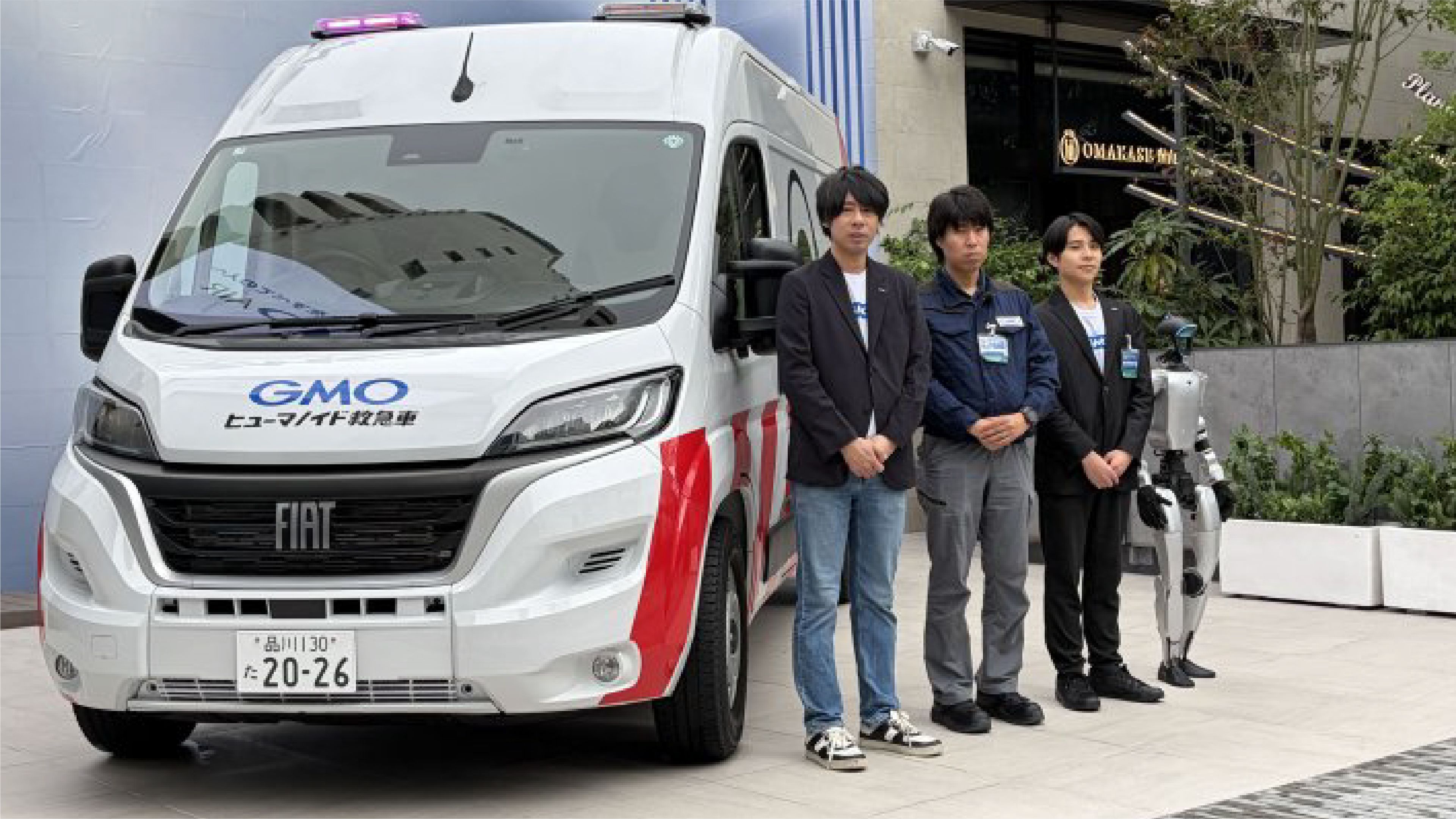Three GMO employees stand beside a white humanoid robot repair ambulance with rooftop lights and two humanoid robots in Tokyo.