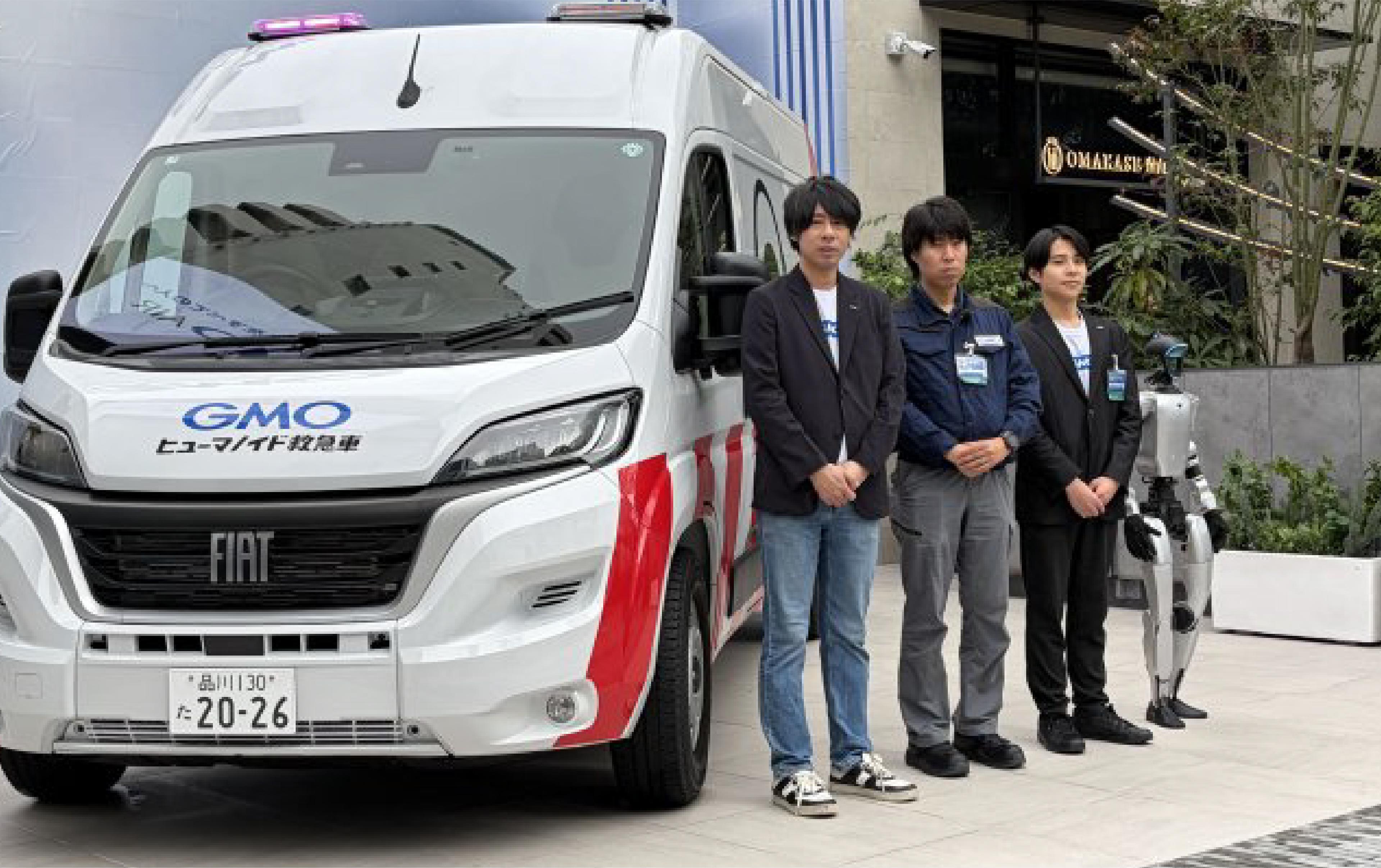 Three GMO employees stand beside a white humanoid robot repair ambulance with rooftop lights and two humanoid robots in Tokyo.