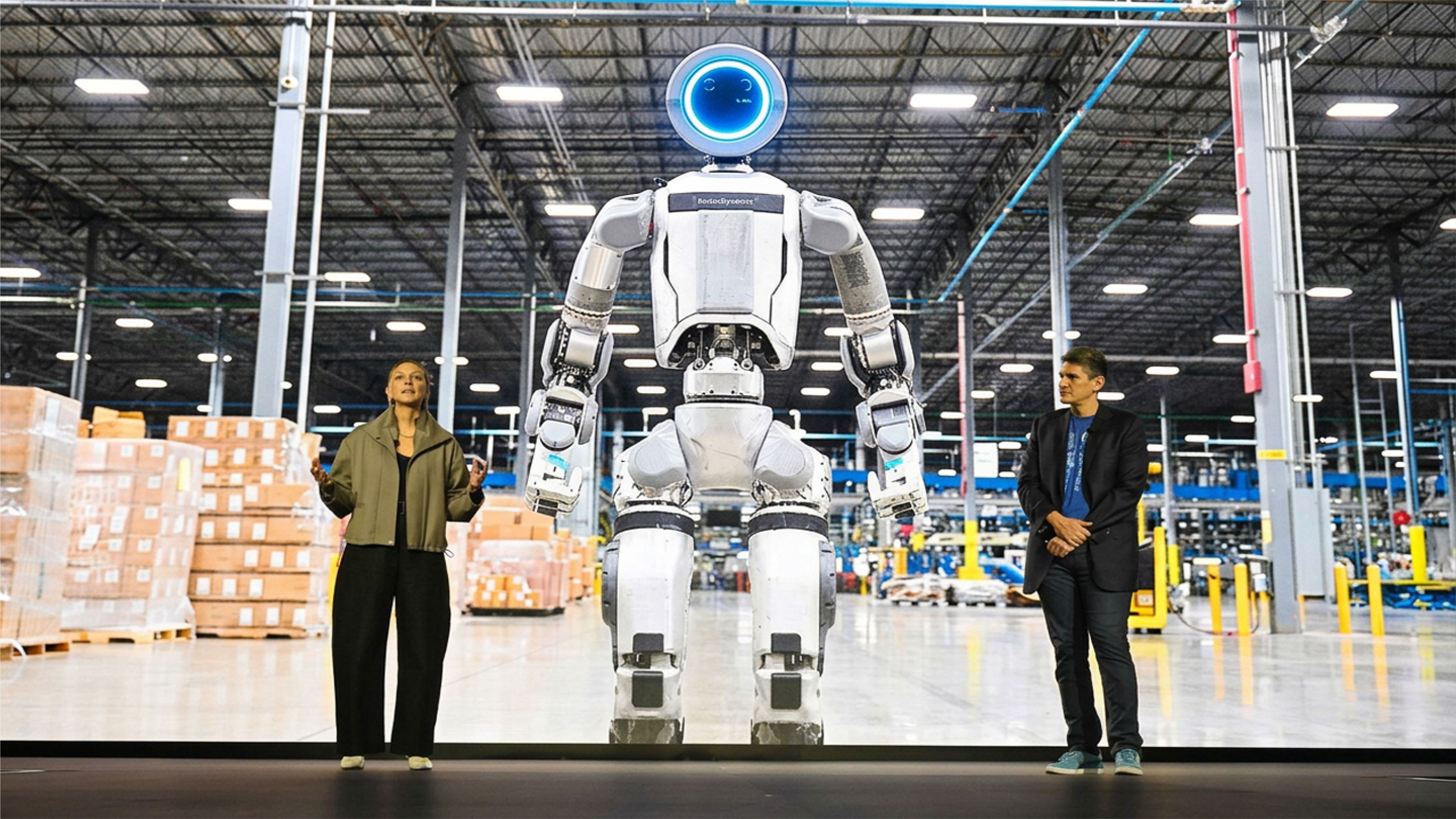 A tall white-and-silver humanoid robot with a glowing blue ring "face" stands on a stage screen inside a large warehouse, its arms hanging at its sides.