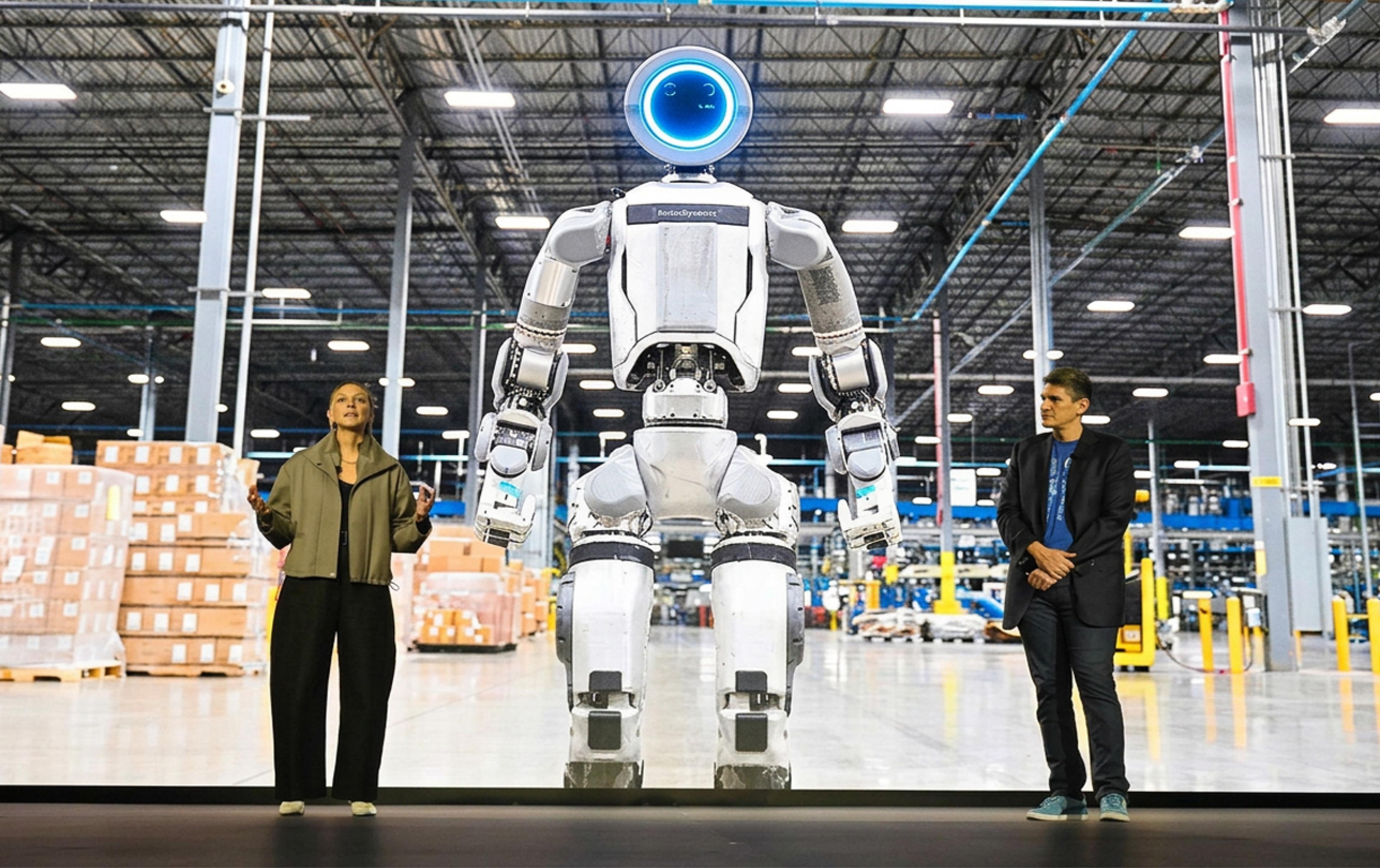 A tall white-and-silver humanoid robot with a glowing blue ring "face" stands on a stage screen inside a large warehouse, its arms hanging at its sides.