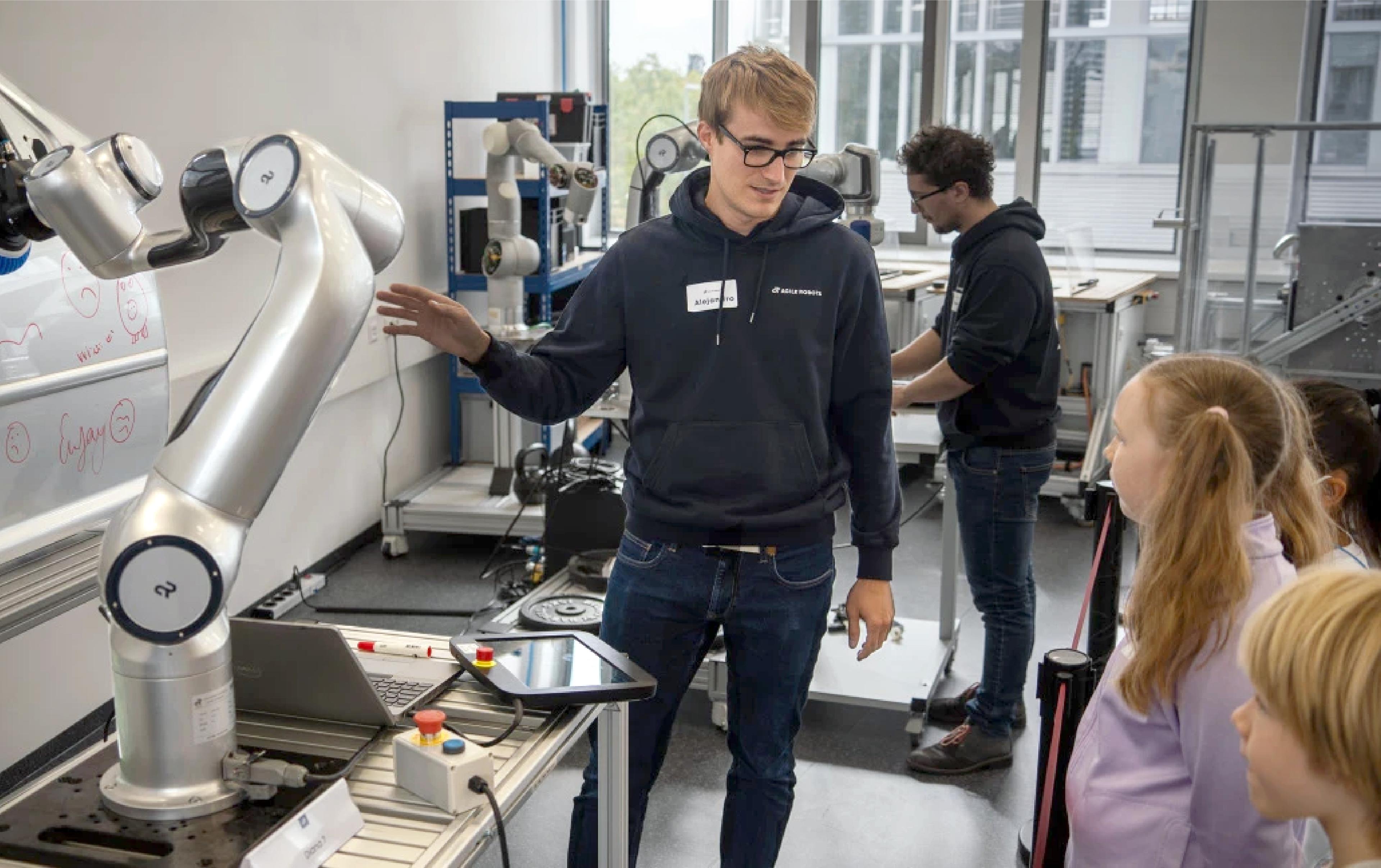 "A young man wearing glasses and a dark hoodie labeled 'Agile Robots' gestures while explaining a silver robotic arm to a small group of children in a lab. The robotic arm, mounted on a workbench with a laptop and control tablet, is drawing on a whiteboard-like panel with a marker. In the background, another person in a similar hoodie works at a second robotic arm station near large windows."