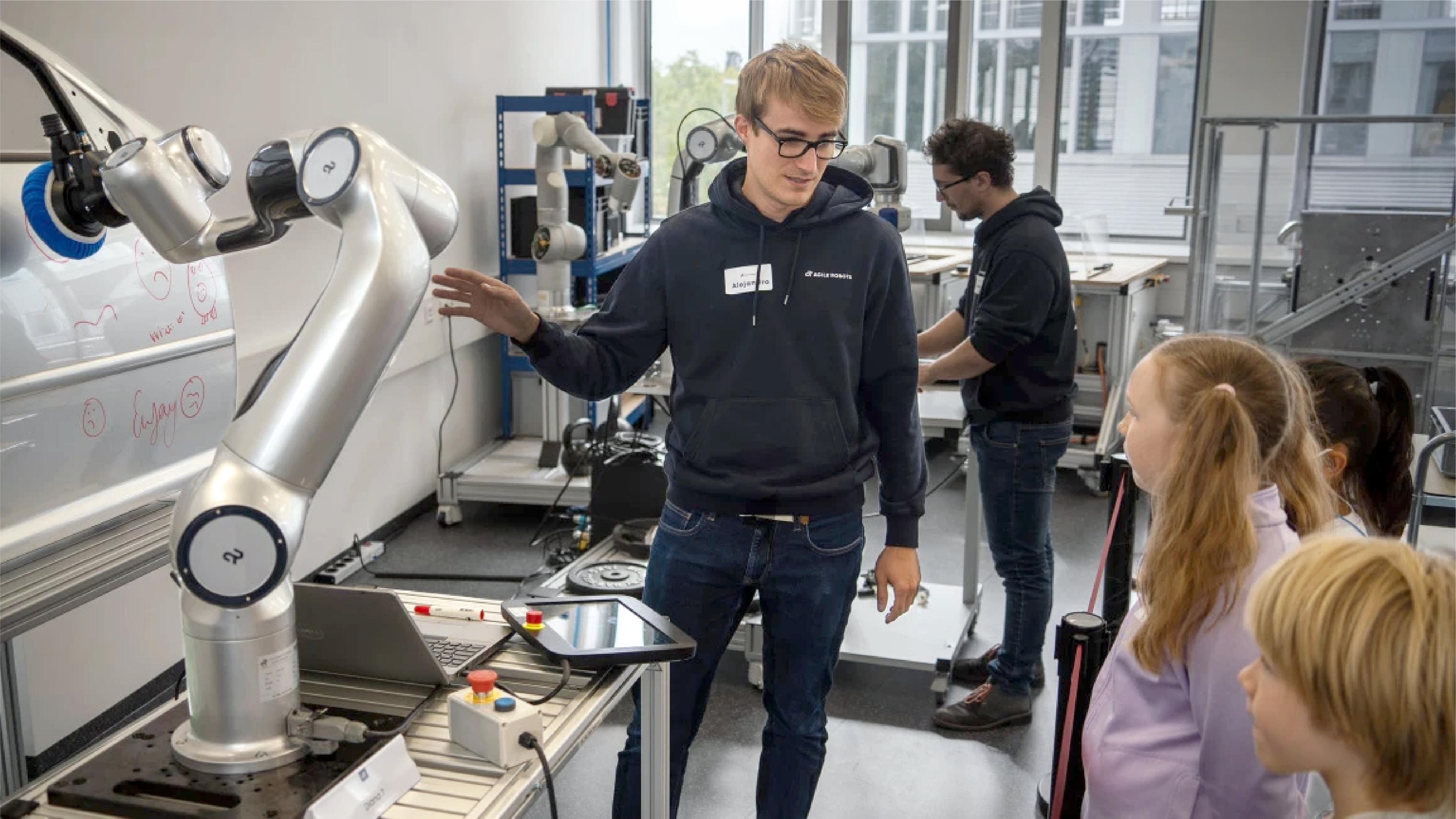 "A young man wearing glasses and a dark hoodie labeled 'Agile Robots' gestures while explaining a silver robotic arm to a small group of children in a lab. The robotic arm, mounted on a workbench with a laptop and control tablet, is drawing on a whiteboard-like panel with a marker. In the background, another person in a similar hoodie works at a second robotic arm station near large windows."