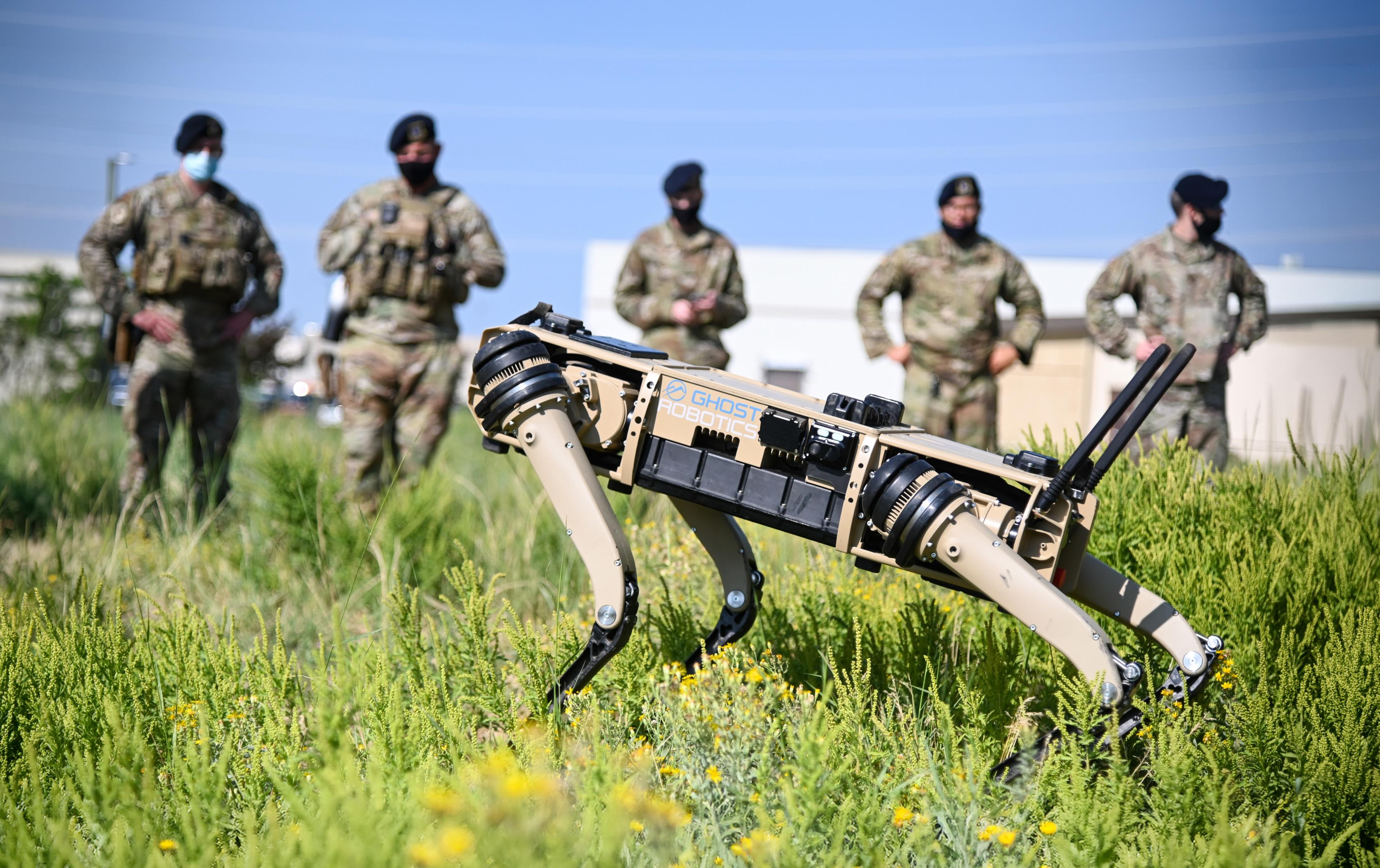 A Ghost Robotics quadruped robot moves through grass during an outdoor military demonstration, with uniformed personnel standing in the background.
