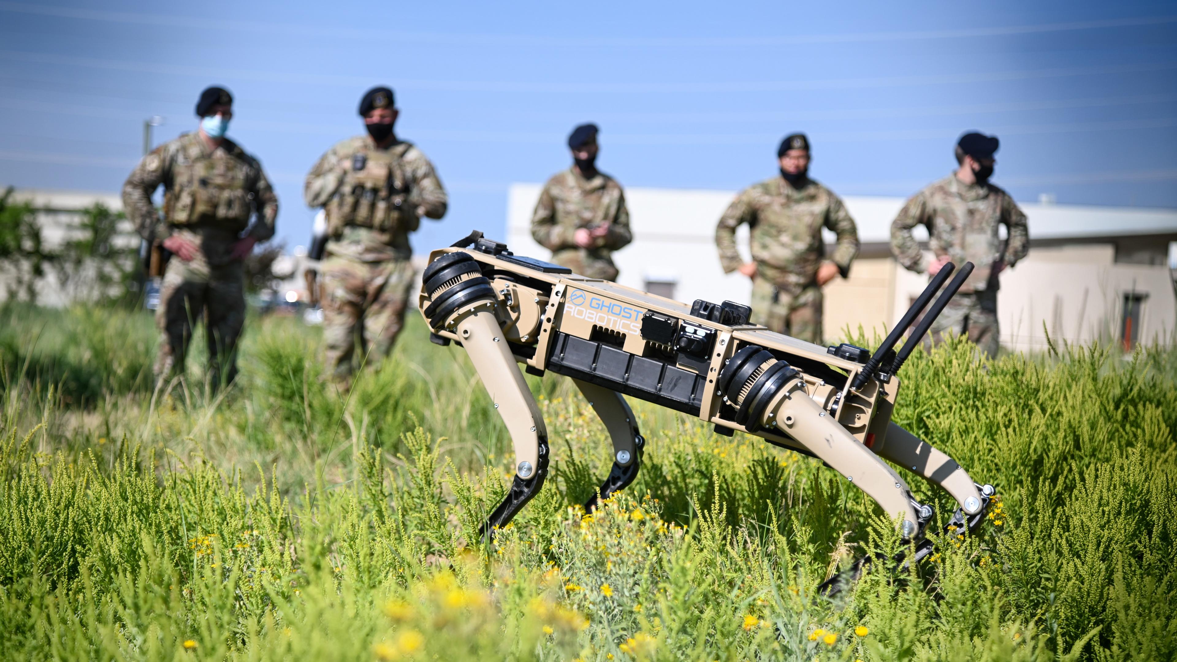 A Ghost Robotics quadruped robot moves through grass during an outdoor military demonstration, with uniformed personnel standing in the background.