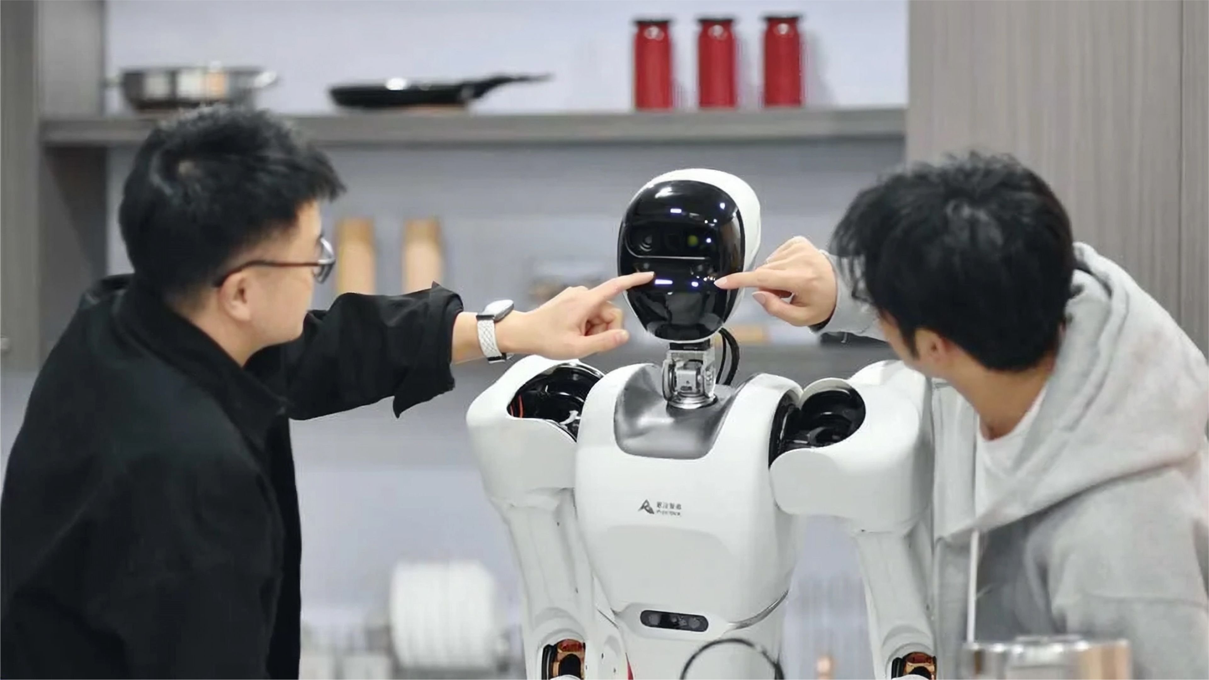 Two people point toward the face of an Astribot humanoid robot with a white body and black sensor-equipped head in a kitchen-like setting.