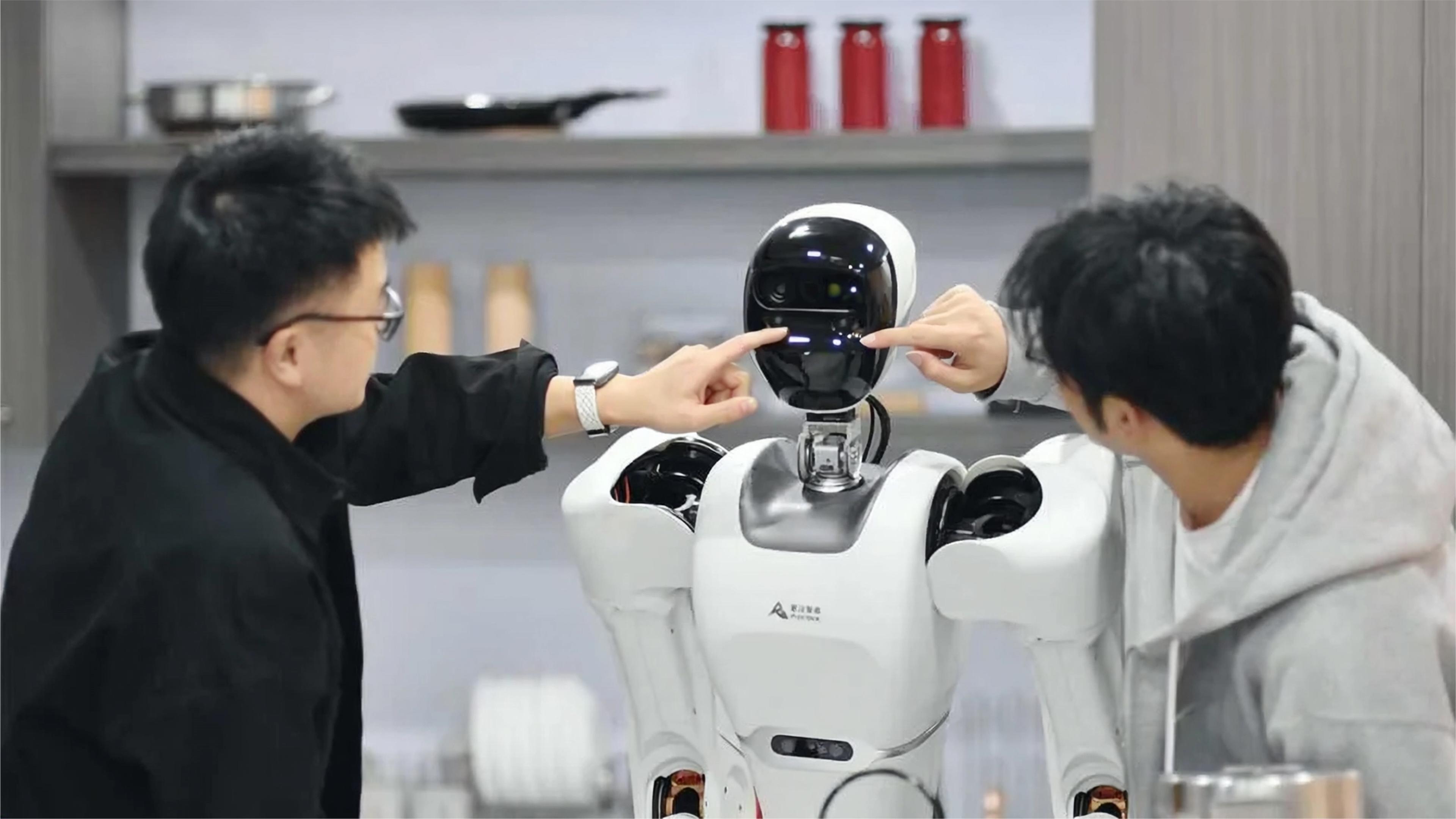 Two people point toward the face of an Astribot humanoid robot with a white body and black sensor-equipped head in a kitchen-like setting.