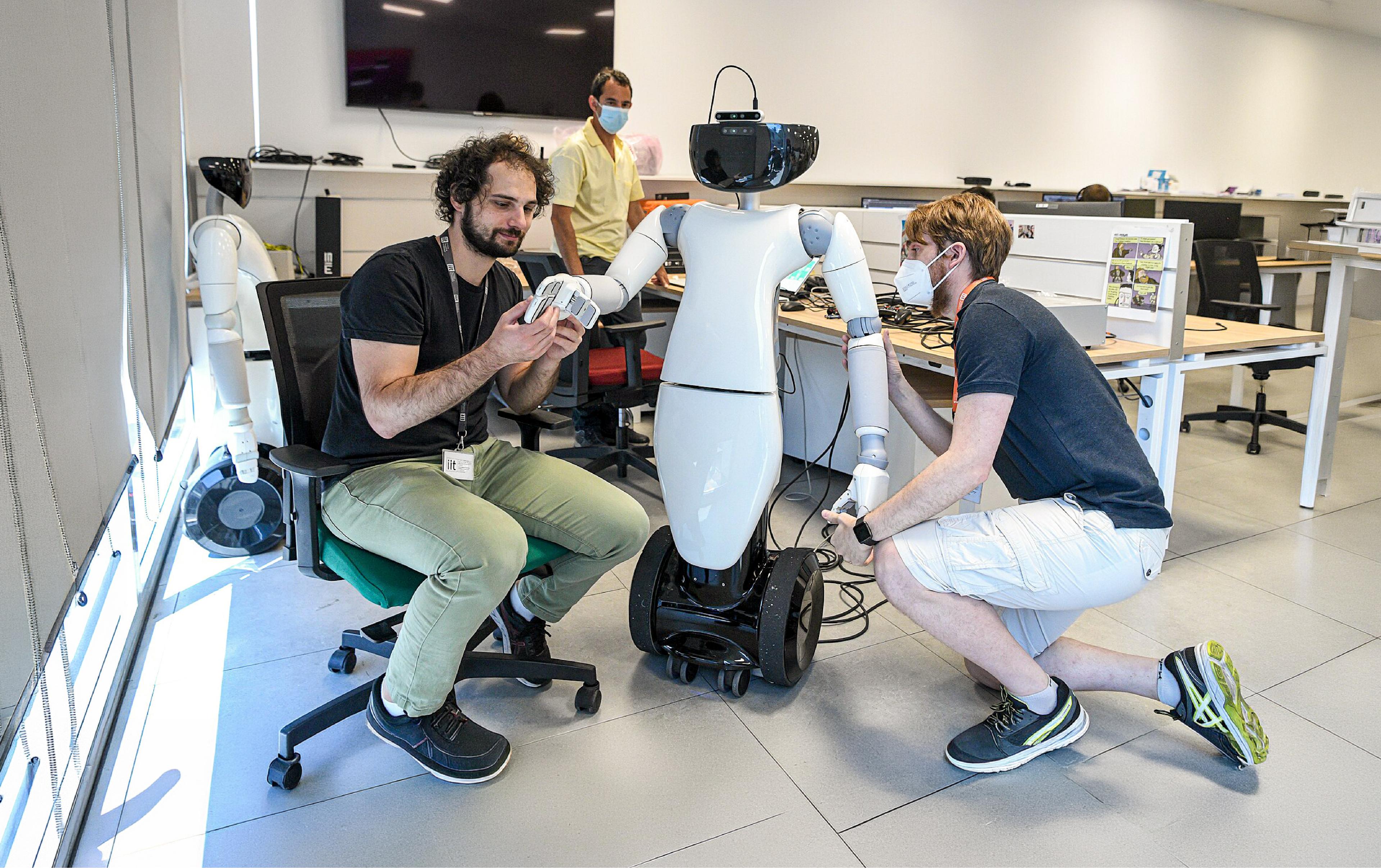 Two researchers adjust the hands and arms of a white humanoid robot on a wheeled base in a robotics laboratory.