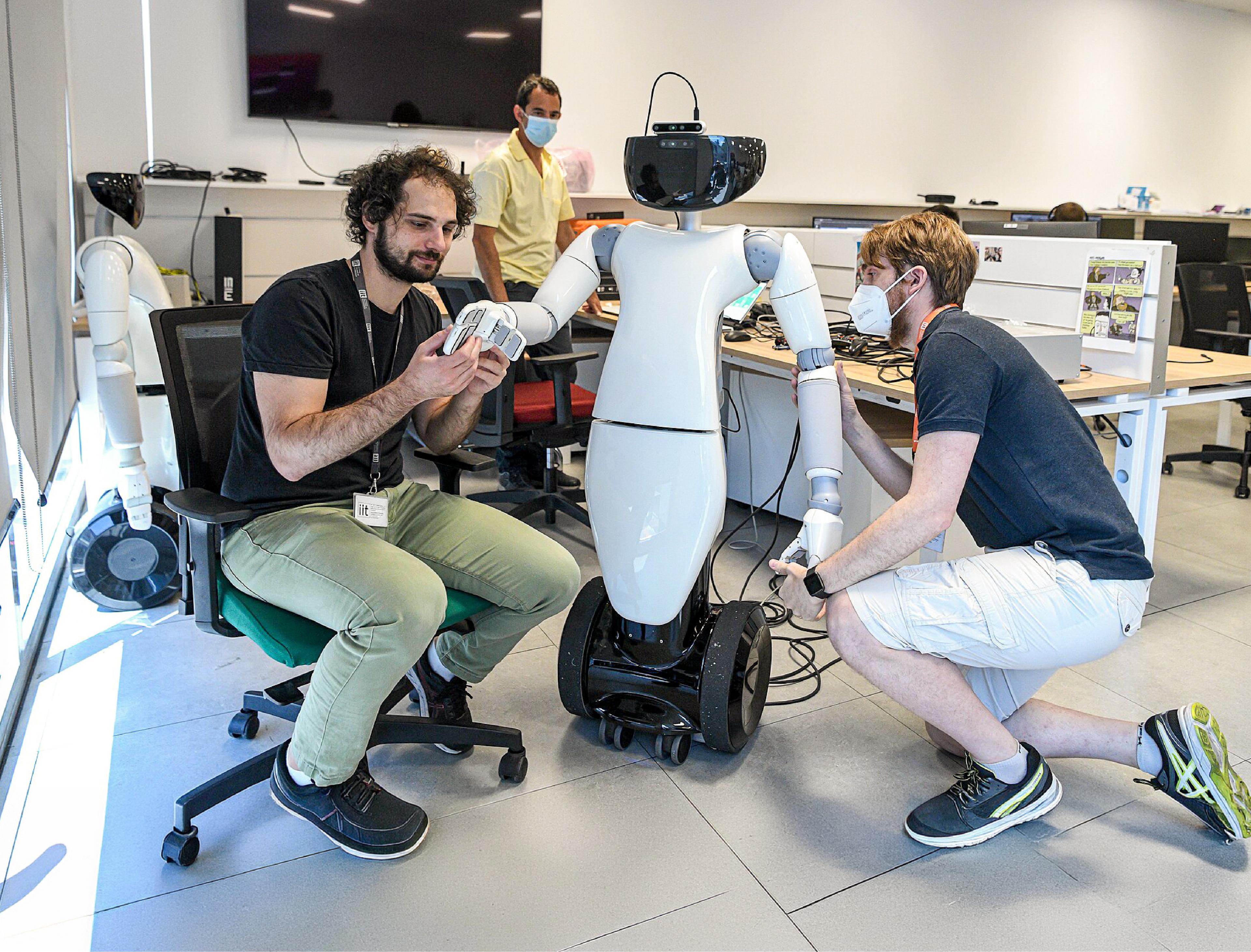 Two researchers adjust the hands and arms of a white humanoid robot on a wheeled base in a robotics laboratory.