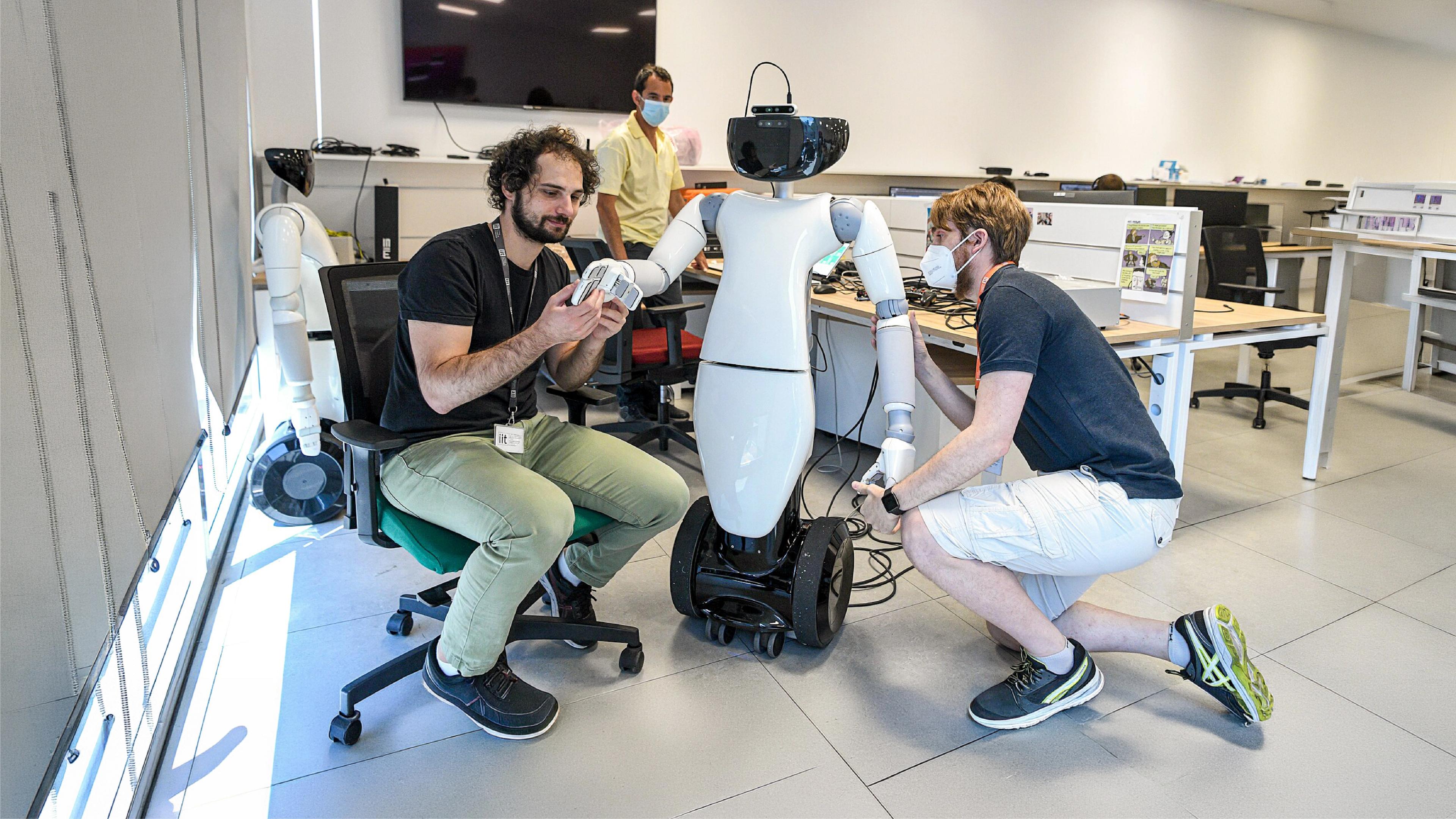 Two researchers adjust the hands and arms of a white humanoid robot on a wheeled base in a robotics laboratory.