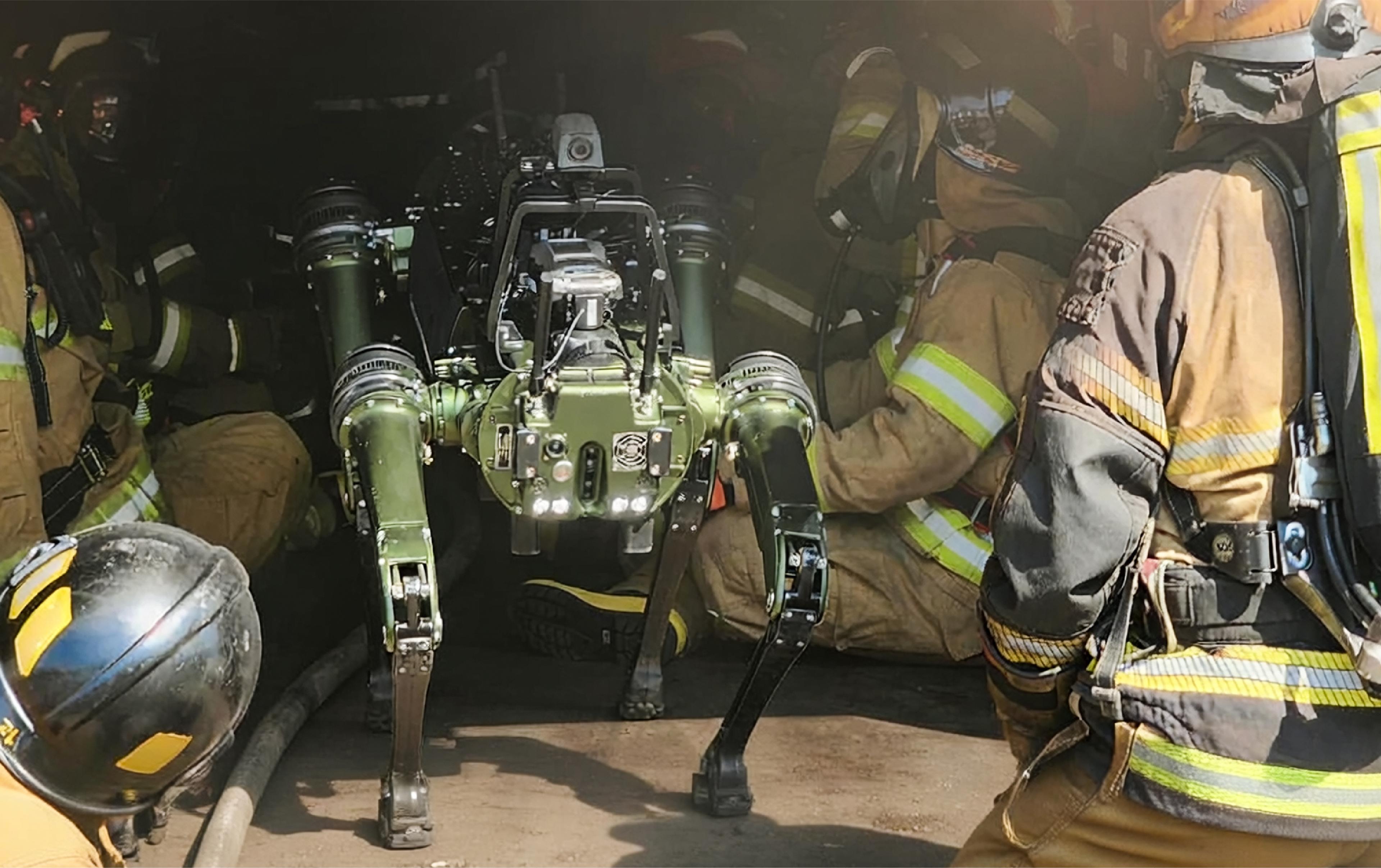 A quadruped robot stands among firefighters during an emergency response training exercise inside a dark, smoke-filled structure.