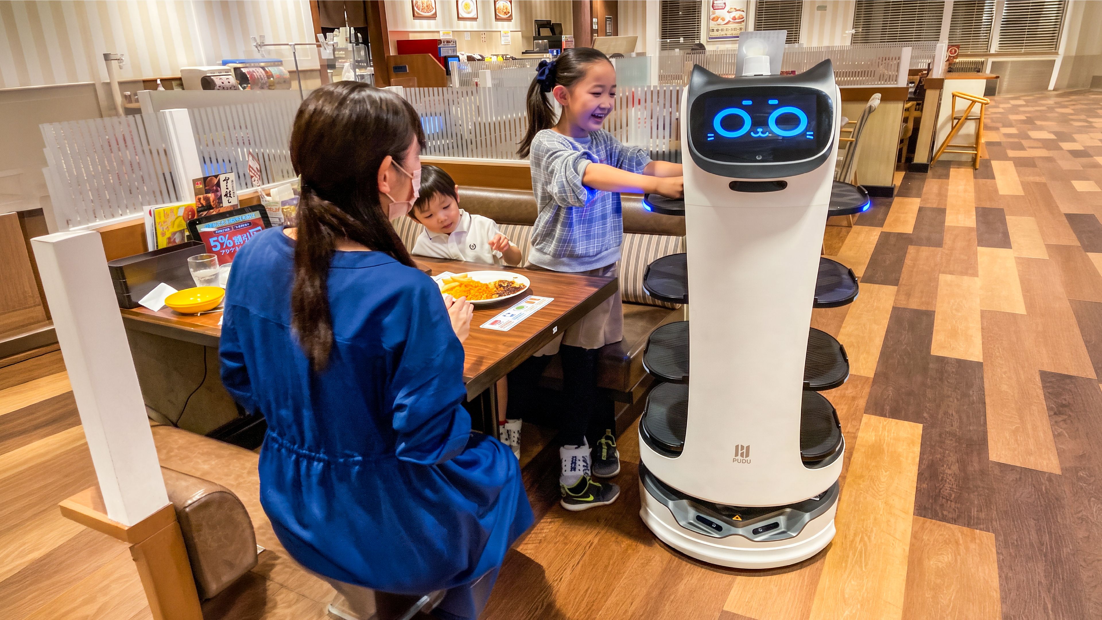 Serving robots deliver plates to diners while a staff member loads a bussing robot