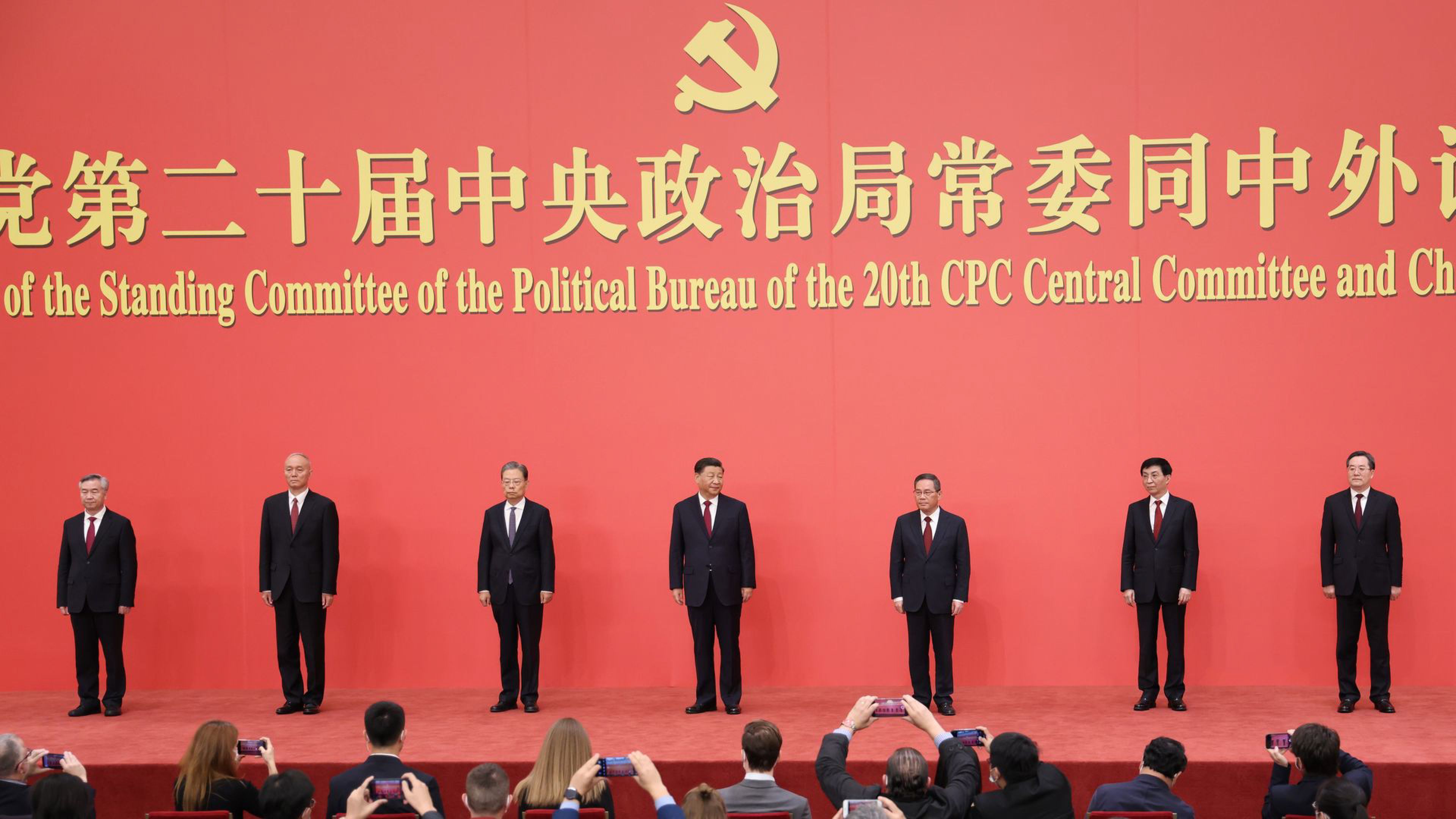 Seven Chinese officials stand beneath the Communist Party emblem at a formal political event as attendees take photos.