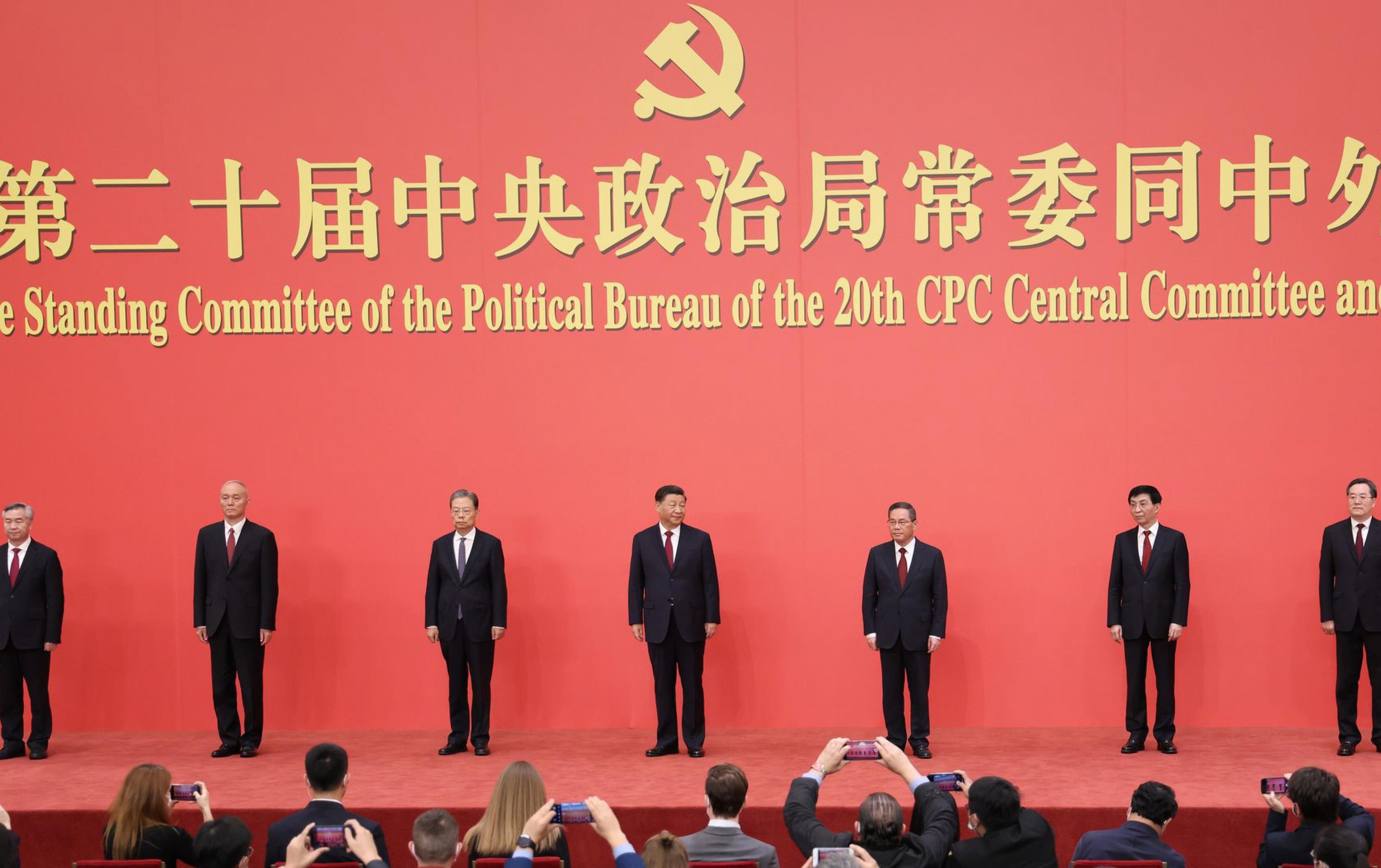 Seven Chinese officials stand beneath the Communist Party emblem at a formal political event as attendees take photos.
