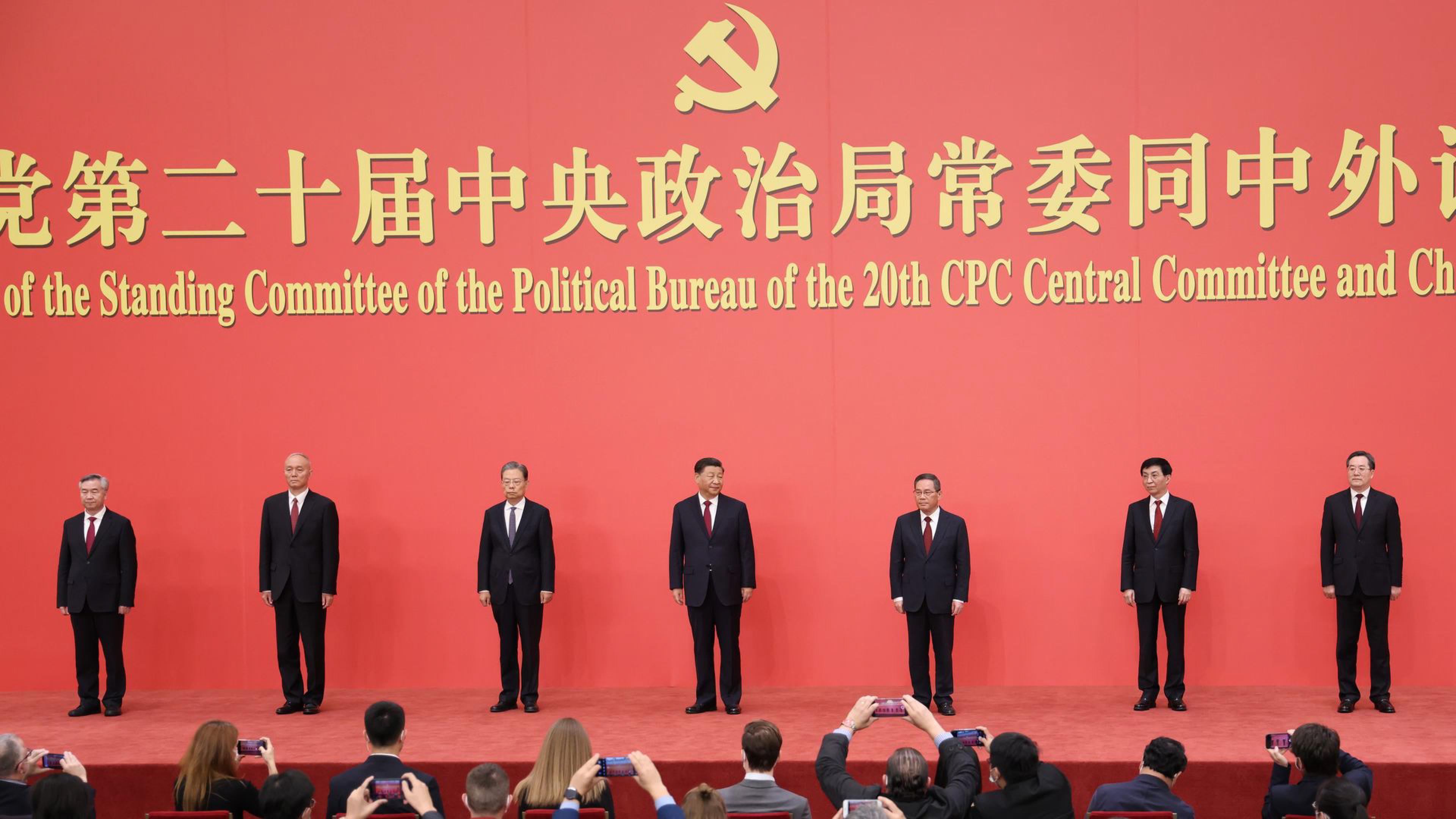 Seven Chinese officials stand beneath the Communist Party emblem at a formal political event as attendees take photos.