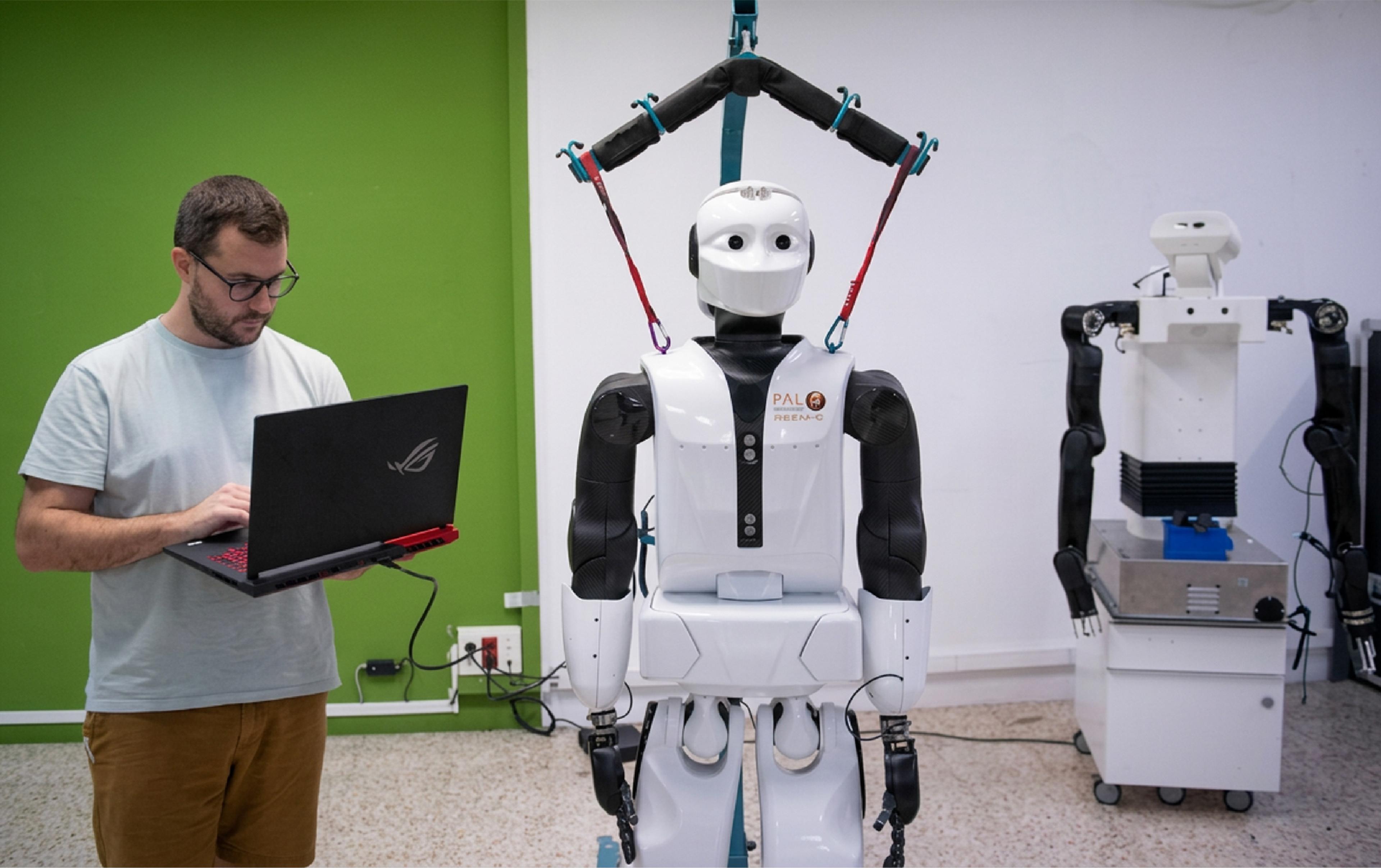 A developer uses a laptop beside a PAL Robotics humanoid robot in a robotics laboratory.