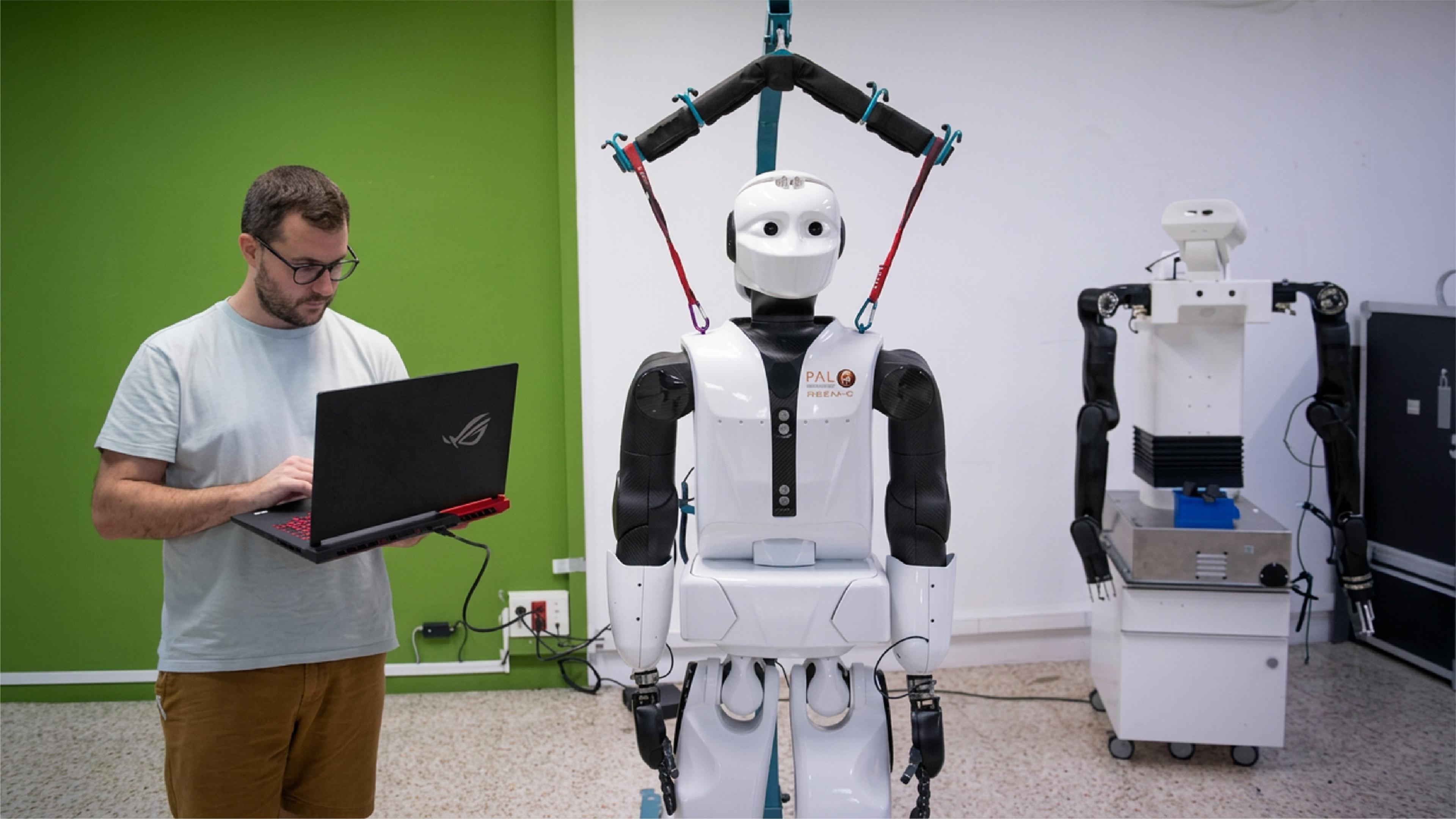A developer uses a laptop beside a PAL Robotics humanoid robot in a robotics laboratory.