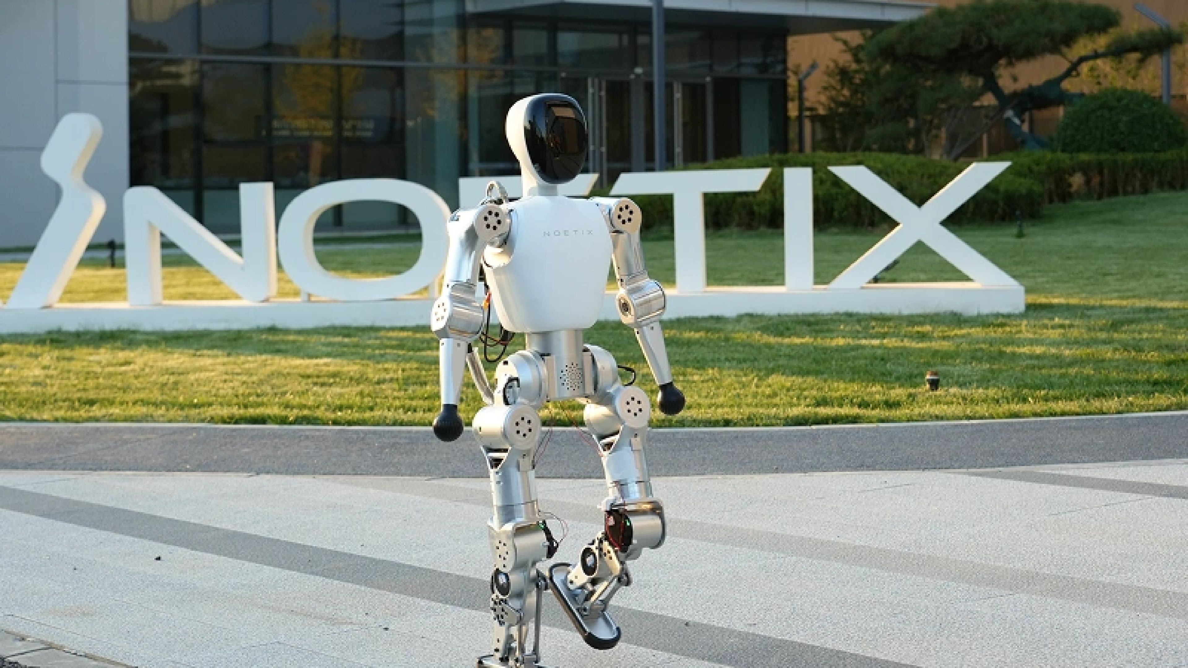 A white Noetix humanoid robot walks outdoors in front of the company's large logo and a modern office building.