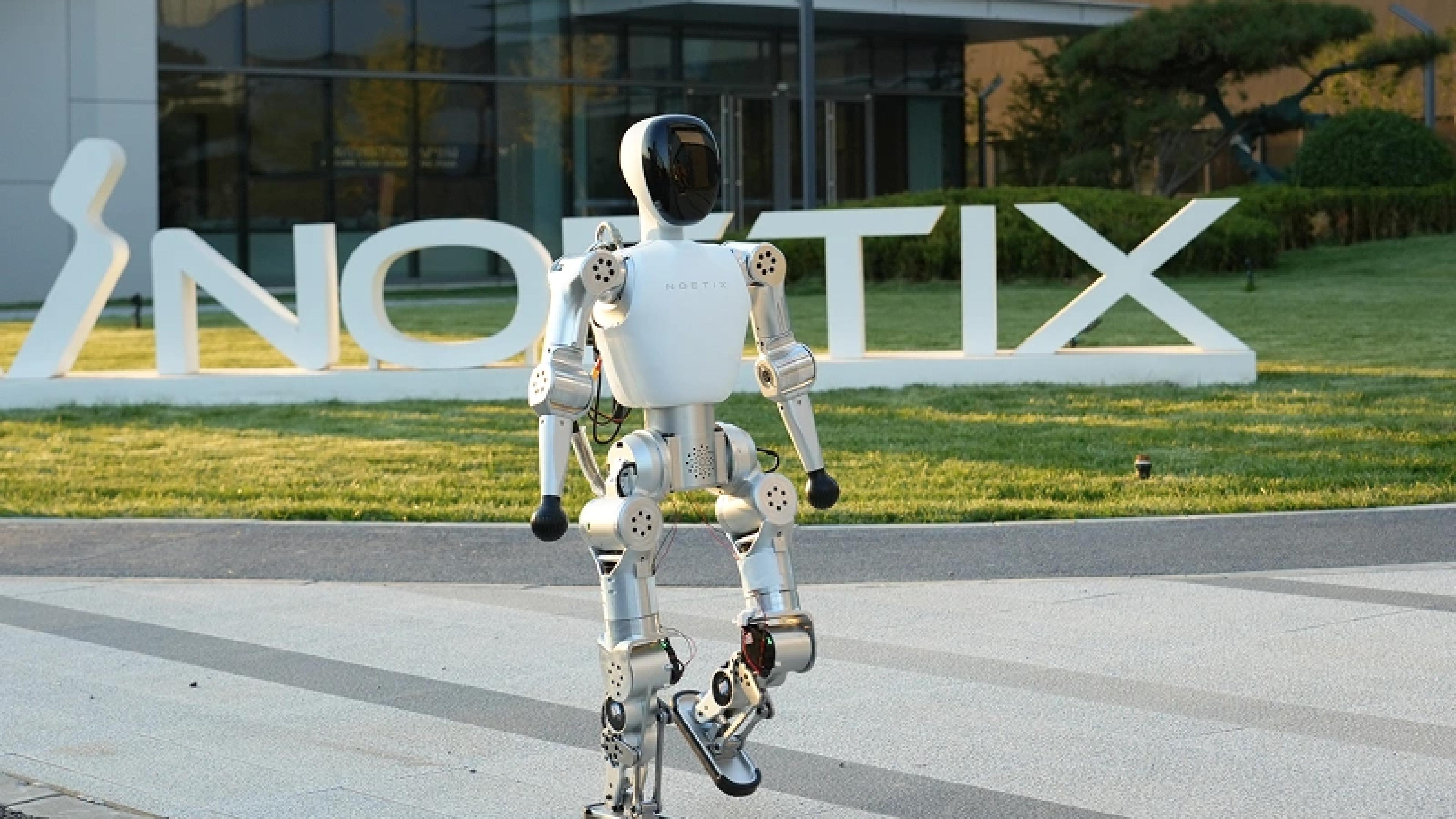 A white Noetix humanoid robot walks outdoors in front of the company's large logo and a modern office building.