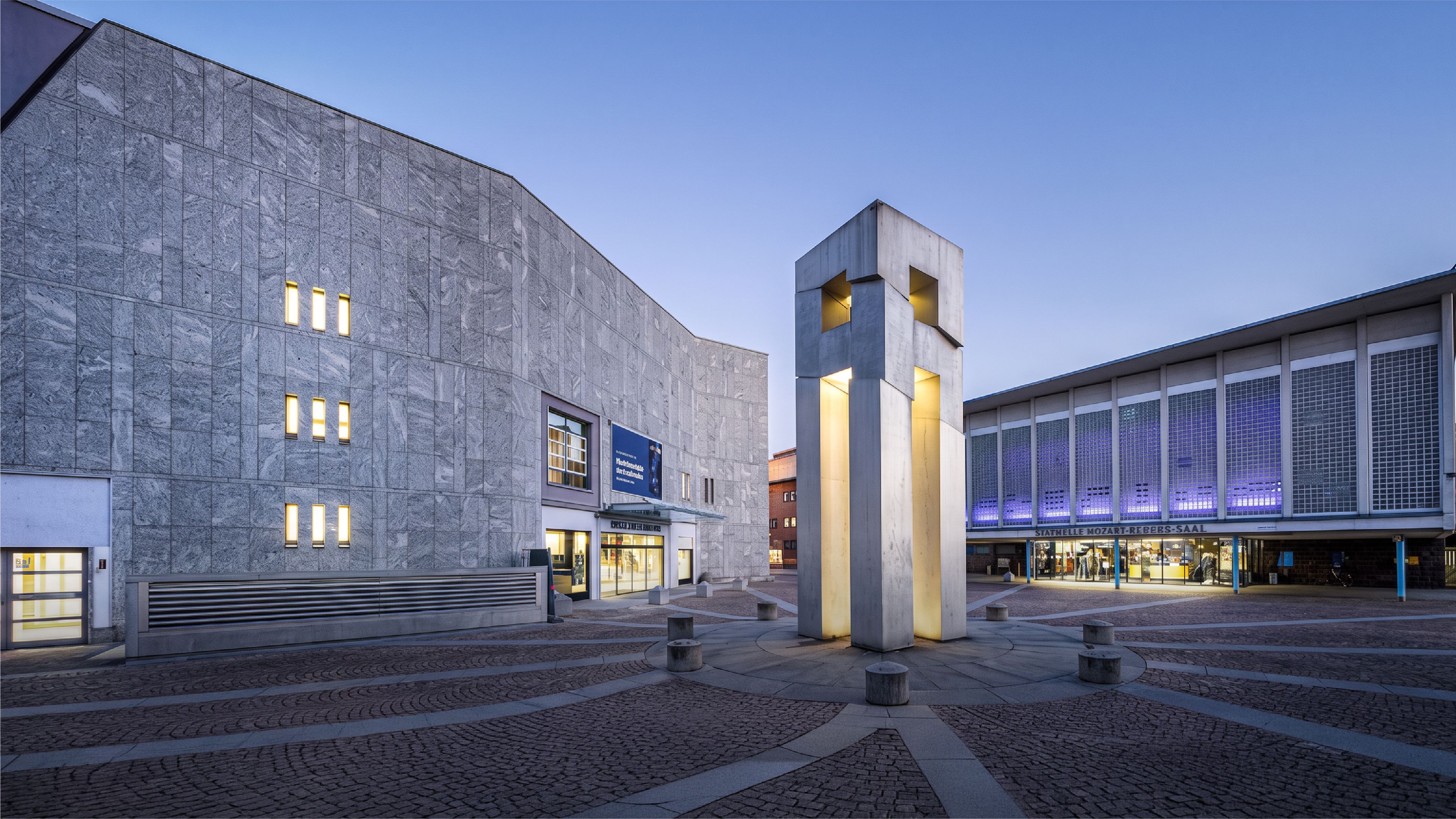 Twilight view of a modern concrete plaza with a stone-clad concert hall on the left, an illuminated sculptural tower in the center, and a glass-fronted building labeled 'Mozart-Rebbes-Saal' on the right, all set on patterned cobblestone paving under a clear blue evening sky."