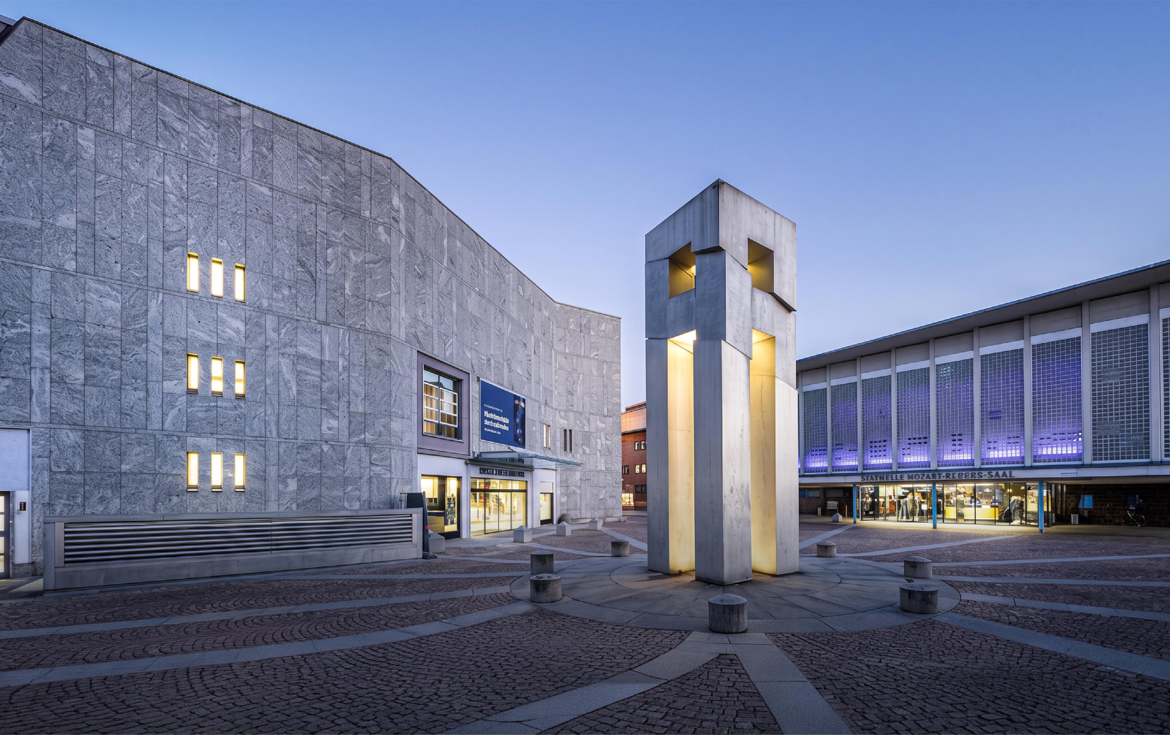 Twilight view of a modern concrete plaza with a stone-clad concert hall on the left, an illuminated sculptural tower in the center, and a glass-fronted building labeled 'Mozart-Rebbes-Saal' on the right, all set on patterned cobblestone paving under a clear blue evening sky."