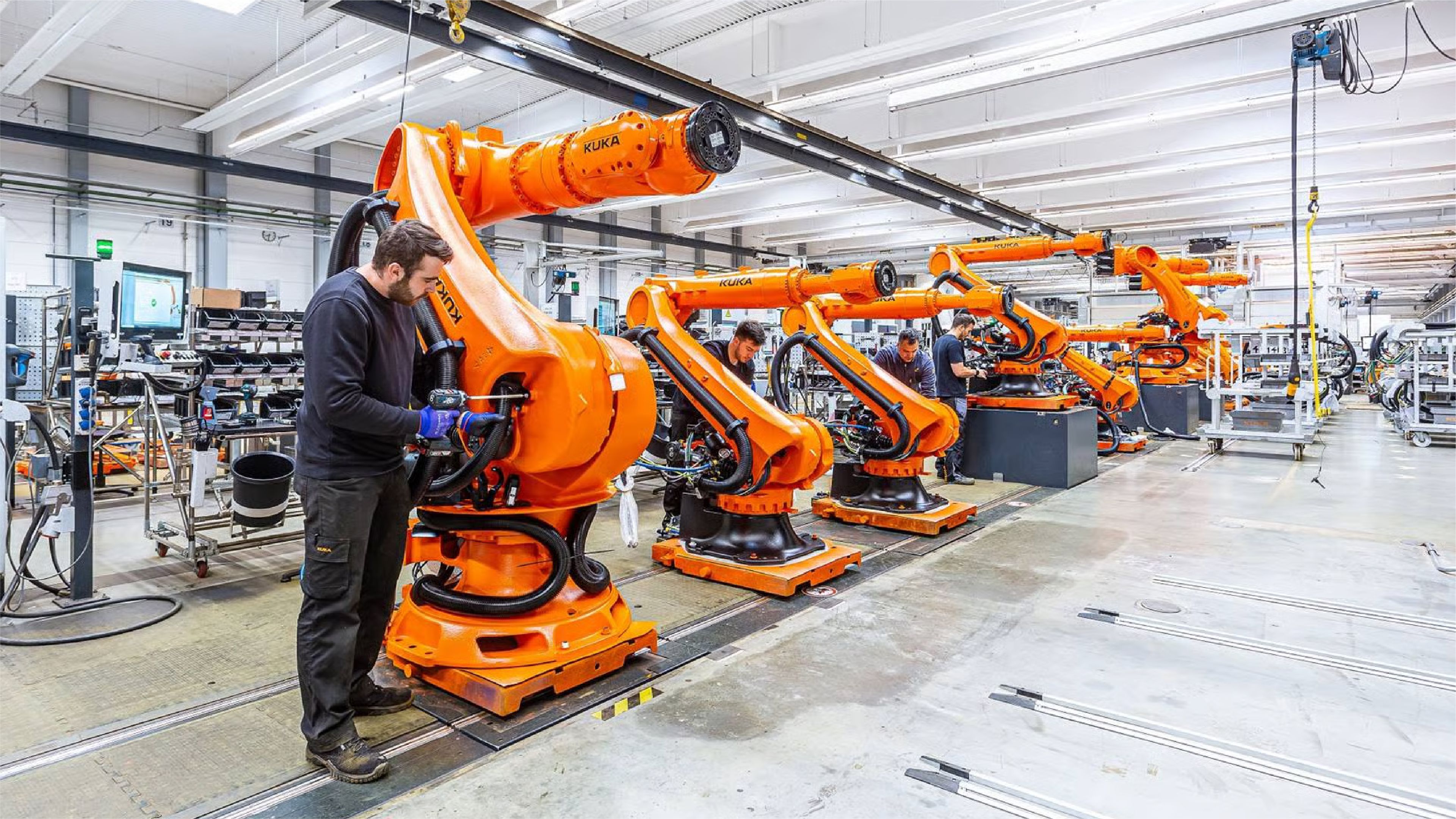 Worker assembling a row of orange KUKA industrial robot arms on a factory production line