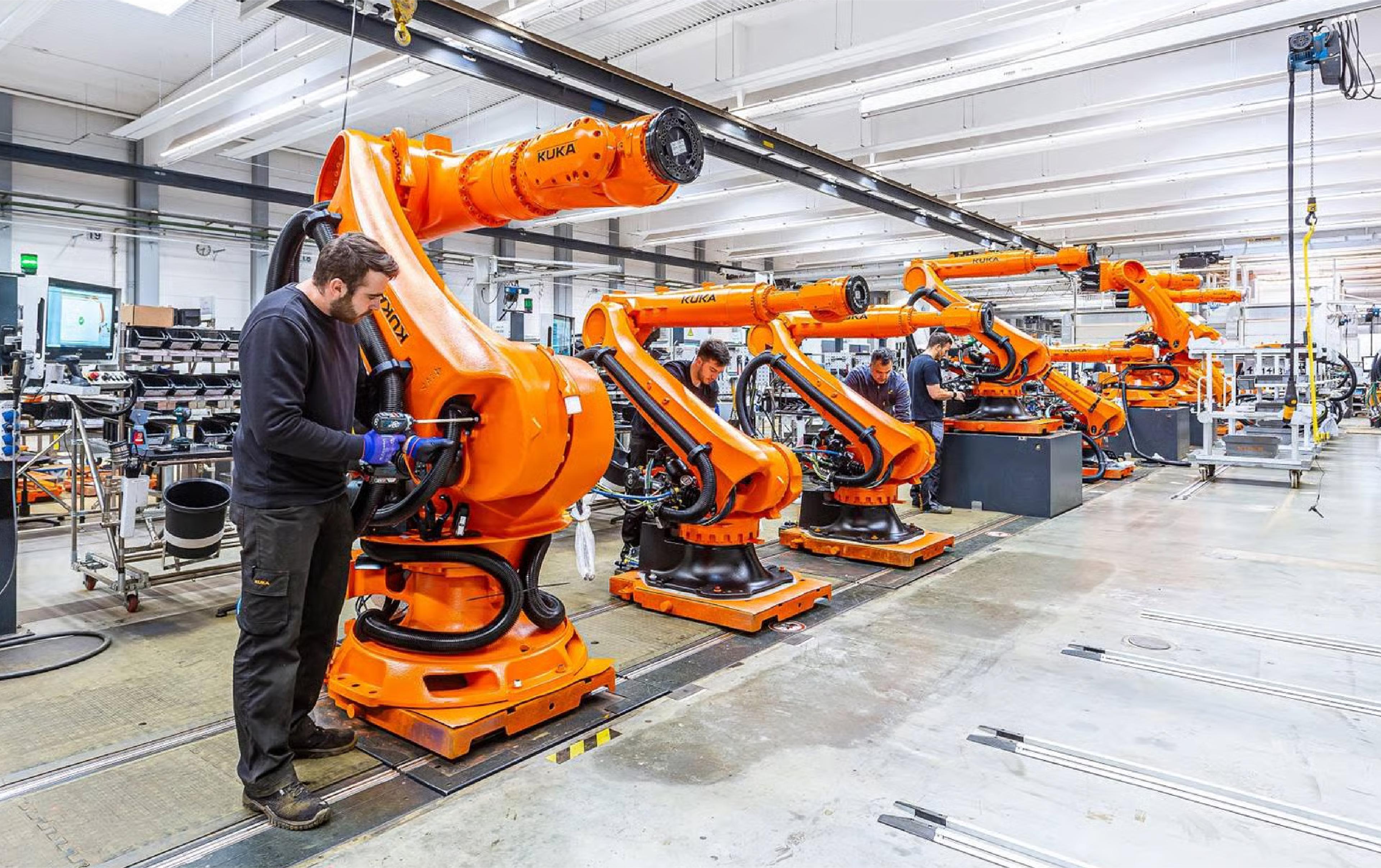 Worker assembling a row of orange KUKA industrial robot arms on a factory production line