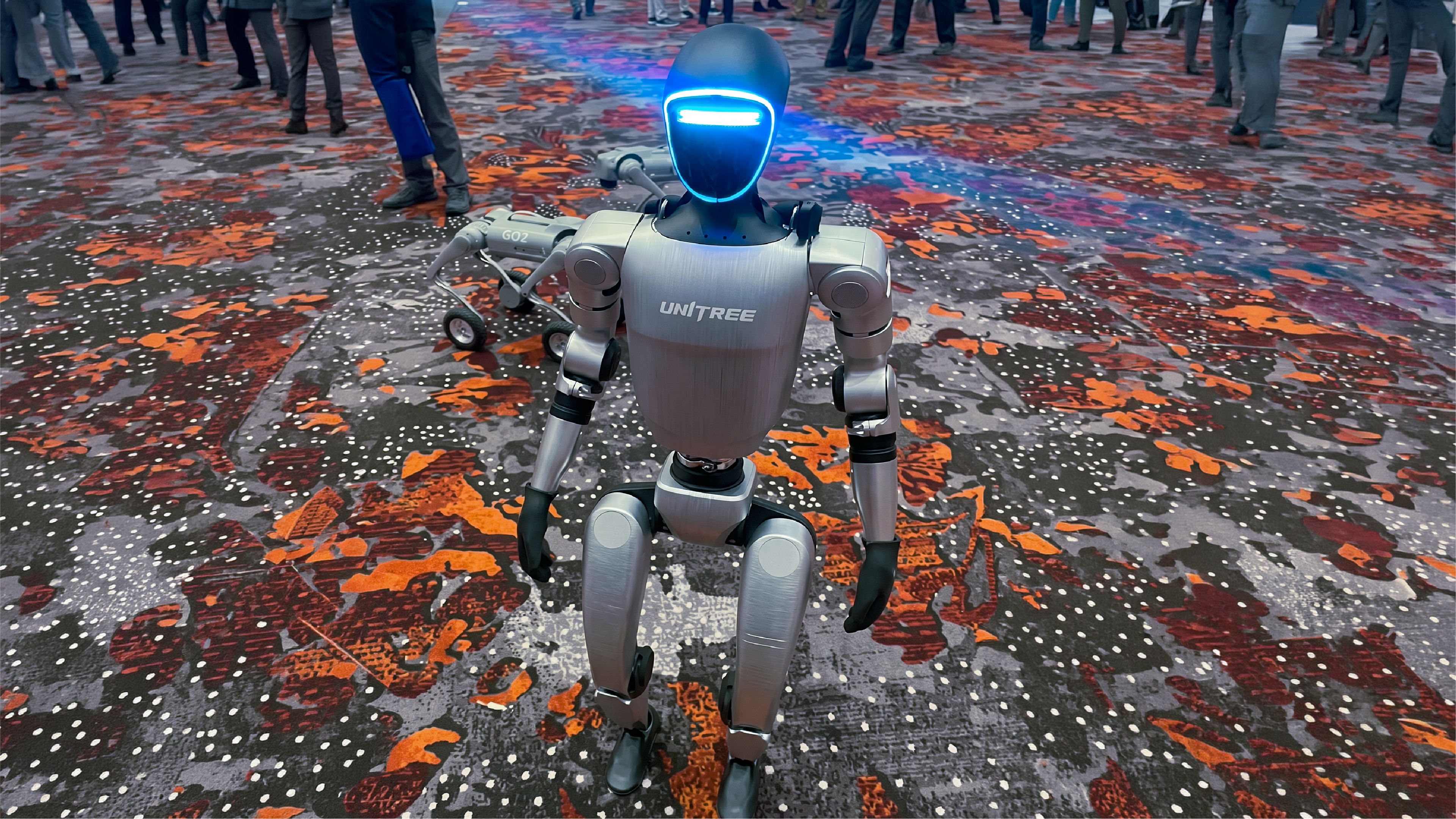 A Unitree humanoid robot with a glowing blue visor stands on a colorful patterned floor at an event, with a Unitree Go2 quadruped robot behind it and a crowd of attendees in the background."