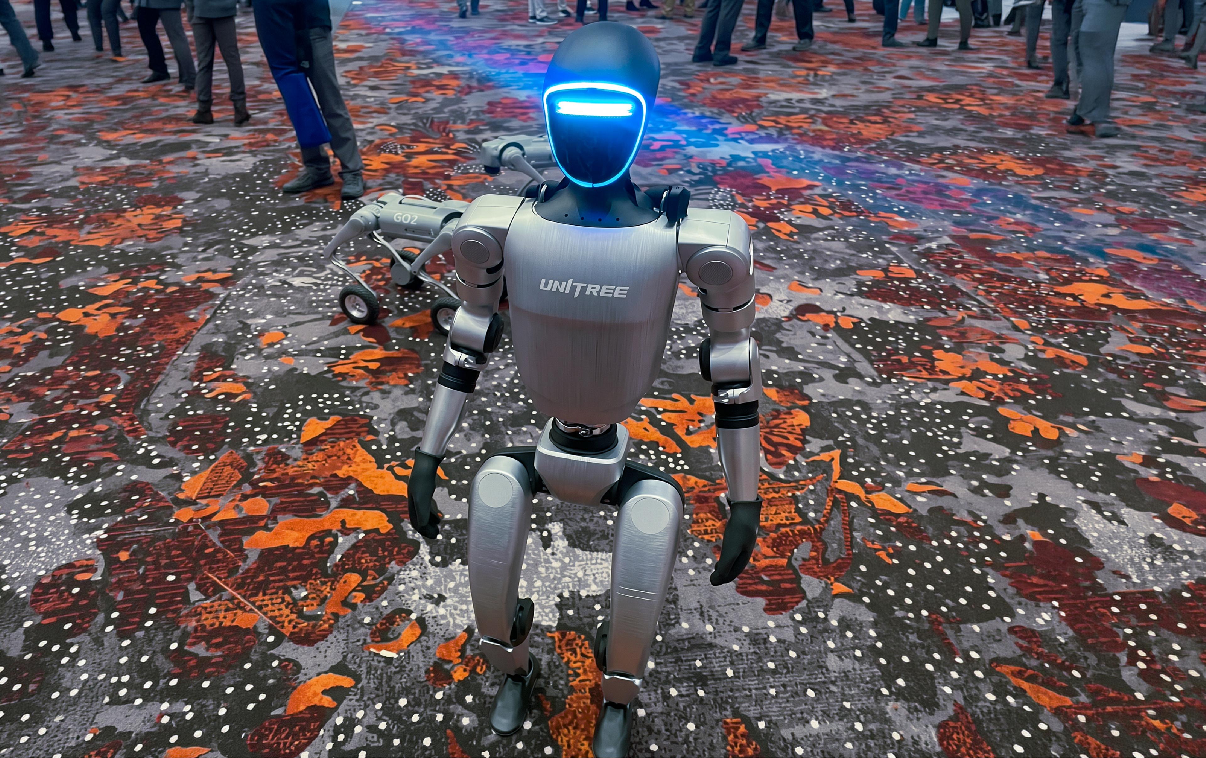 A Unitree humanoid robot with a glowing blue visor stands on a colorful patterned floor at an event, with a Unitree Go2 quadruped robot behind it and a crowd of attendees in the background."