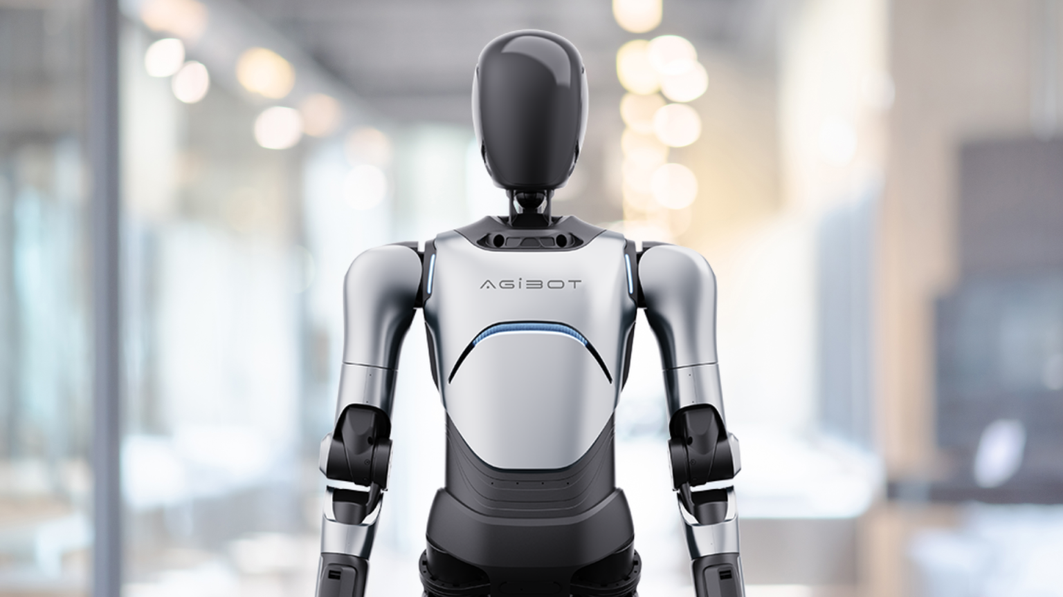 Silver-and-black AgiBot humanoid robot standing in a brightly lit indoor corridor.
