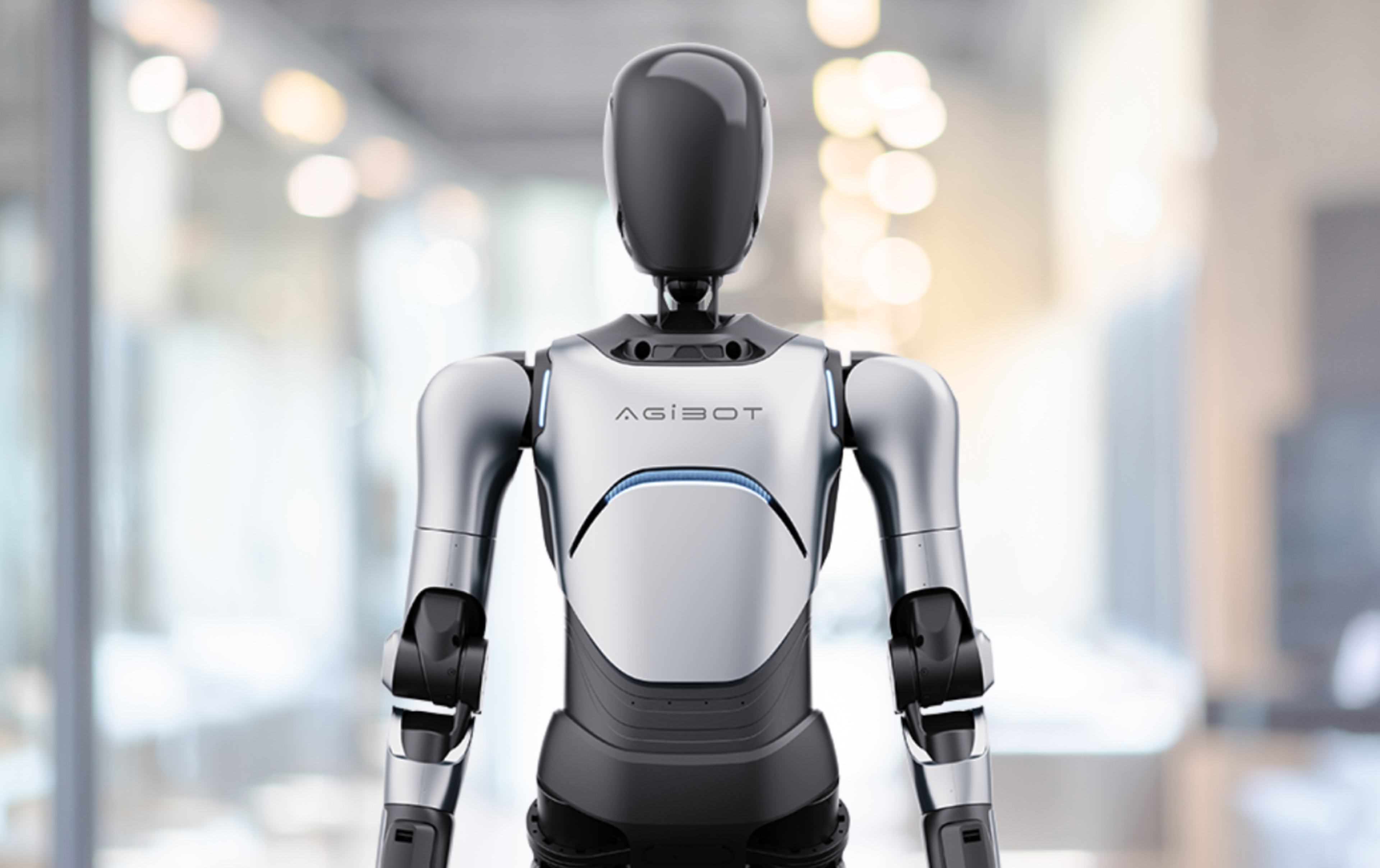Silver-and-black AgiBot humanoid robot standing in a brightly lit indoor corridor.