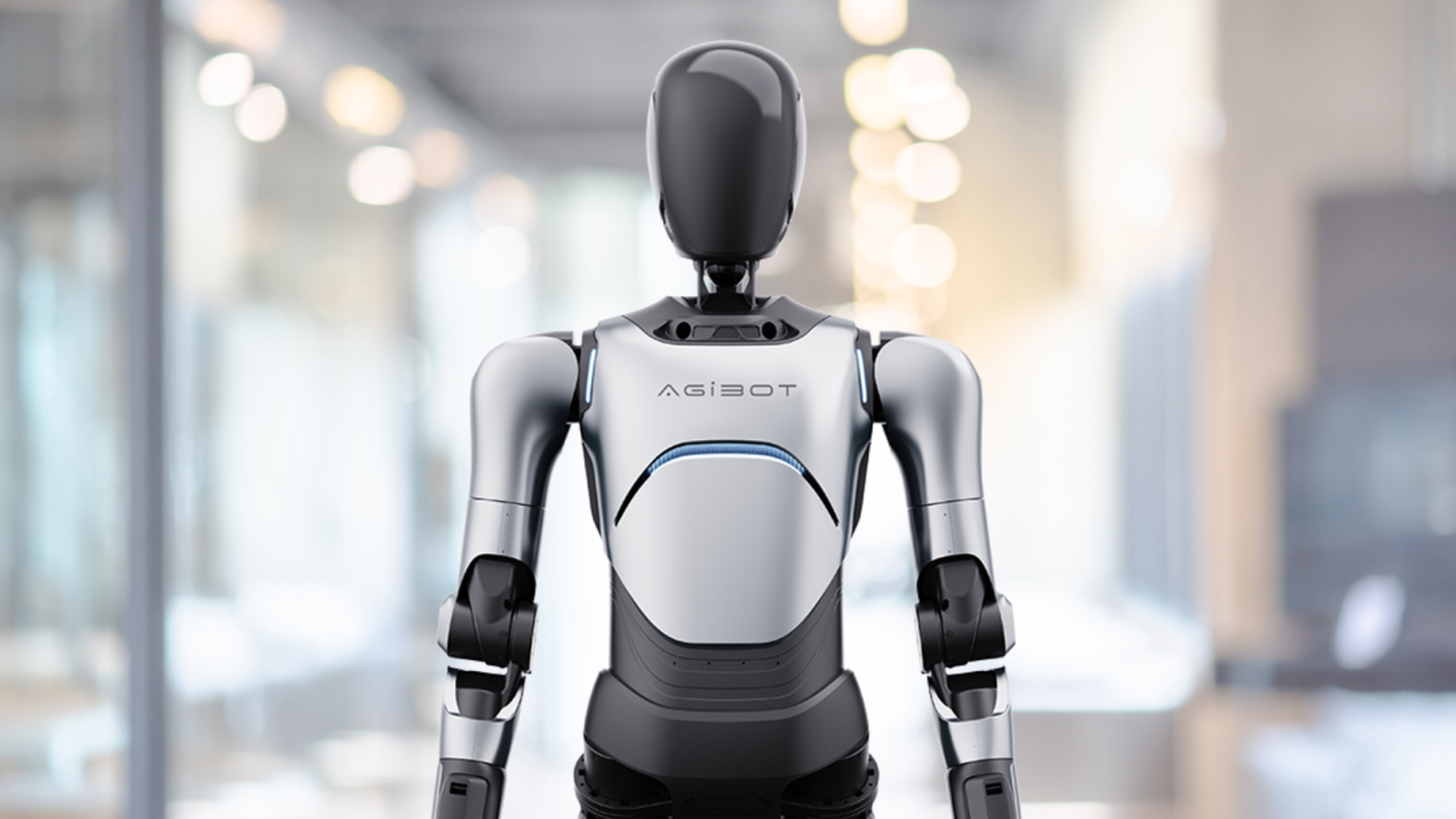 Silver-and-black AgiBot humanoid robot standing in a brightly lit indoor corridor.