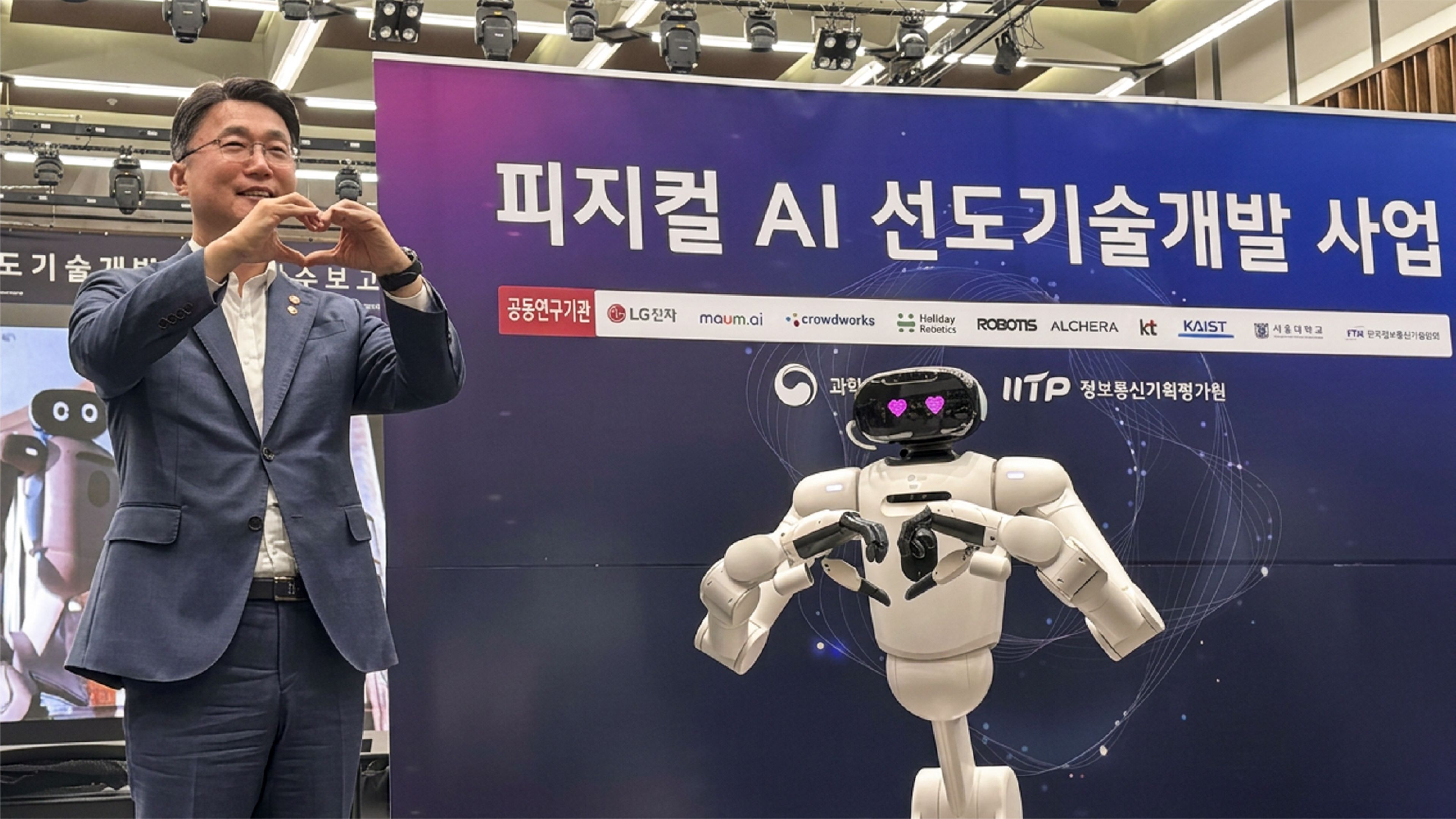 Man in a suit makes a finger-heart gesture beside a white humanoid robot with pink heart eyes at a Korean physical AI event