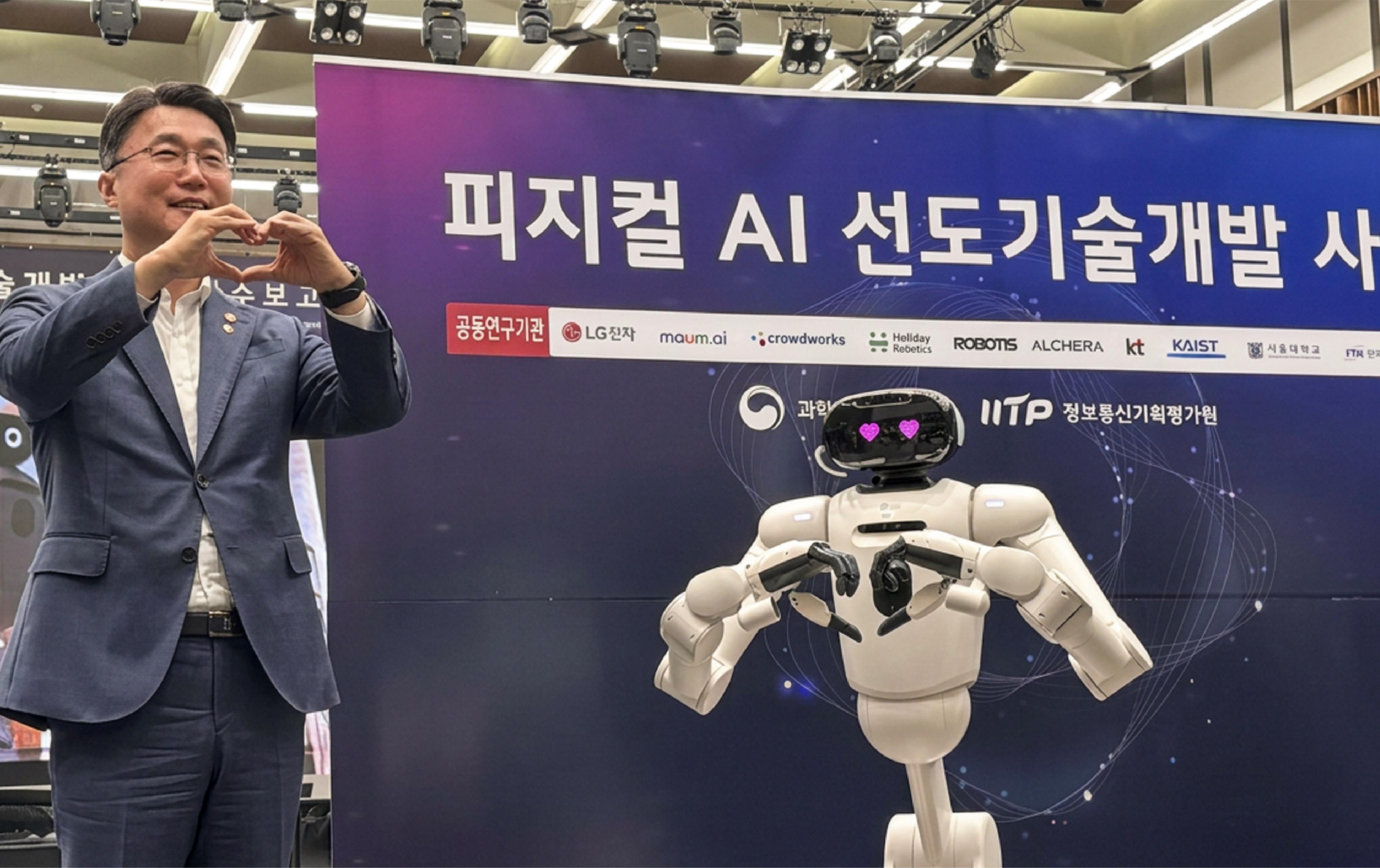 Man in a suit makes a finger-heart gesture beside a white humanoid robot with pink heart eyes at a Korean physical AI event