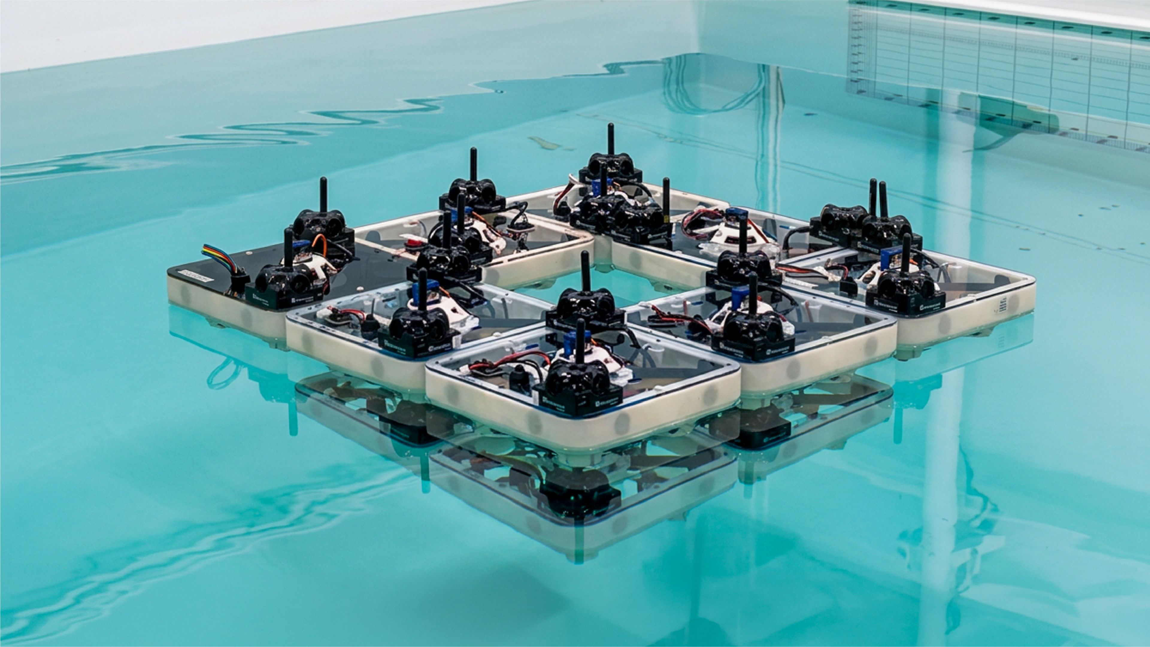 Nine square robotic boats, each with an onboard sensor mast and exposed wiring, latched together edge-to-edge in a 3x3 grid (missing one corner unit) and floating on the still, teal-tinted water of a test tank, with both the robots and the tank's grid-marked wall reflected in the surface.