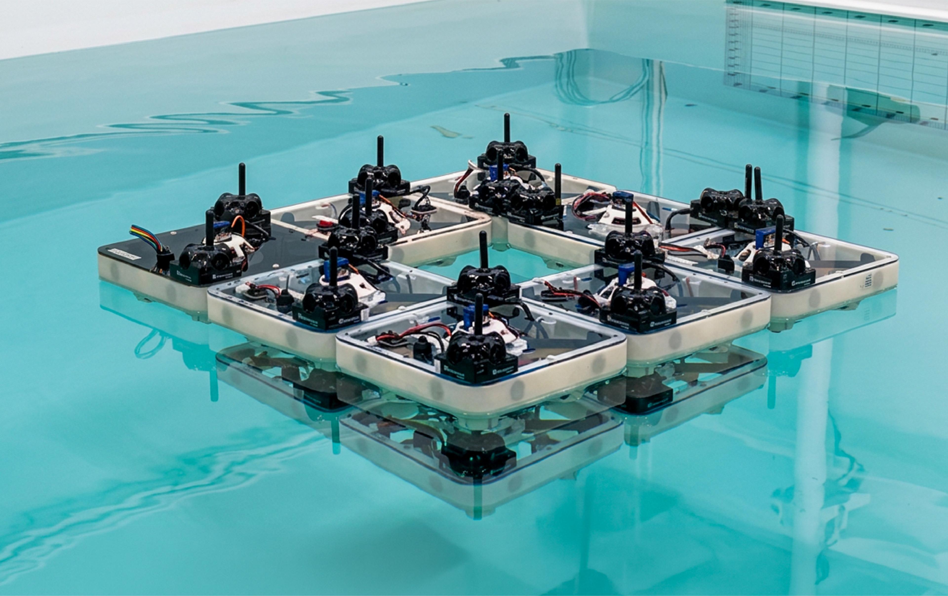 Nine square robotic boats, each with an onboard sensor mast and exposed wiring, latched together edge-to-edge in a 3x3 grid (missing one corner unit) and floating on the still, teal-tinted water of a test tank, with both the robots and the tank's grid-marked wall reflected in the surface.