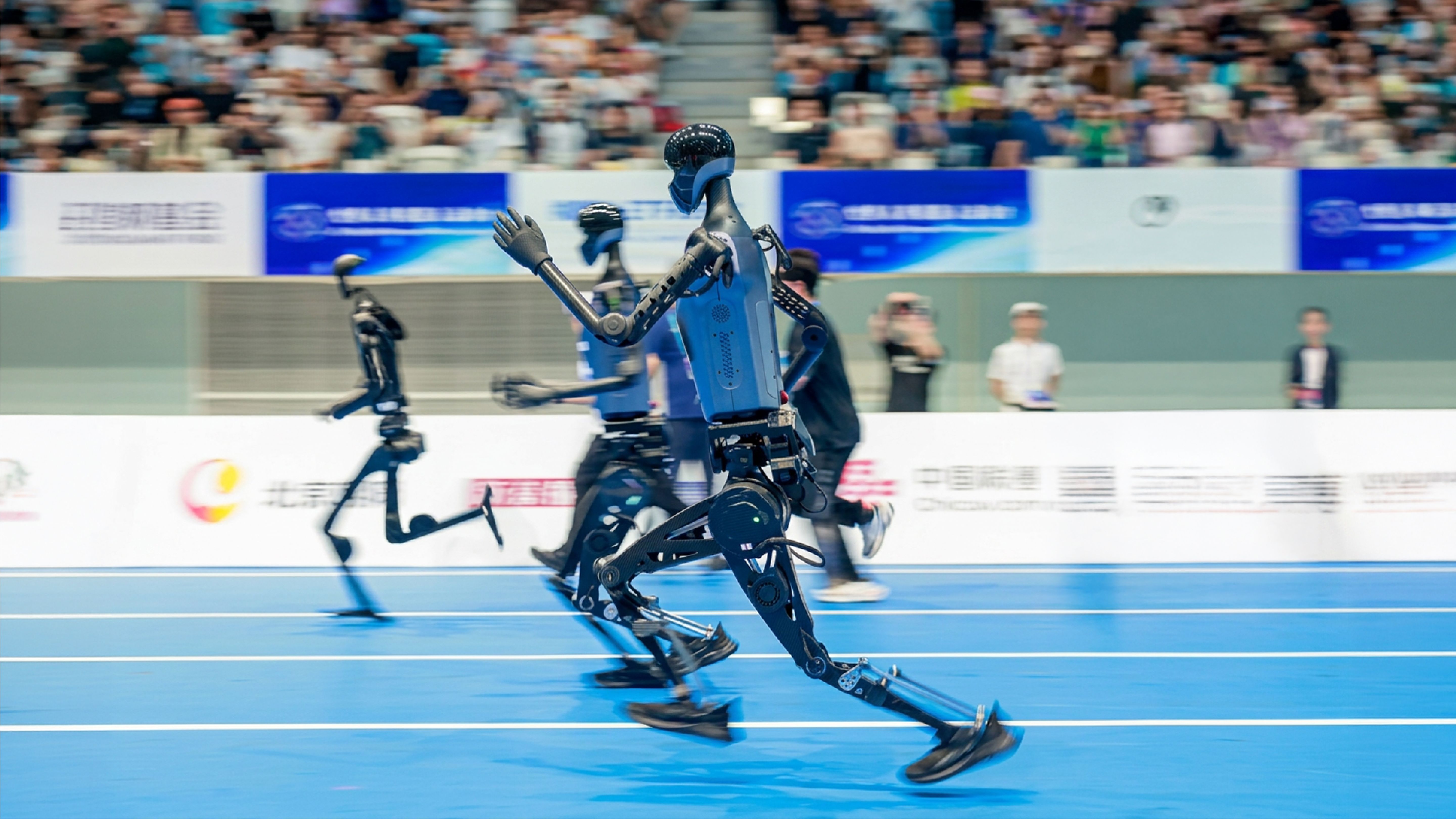 Black humanoid robots sprinting on a blue indoor athletics track in front of a packed stadium crowd