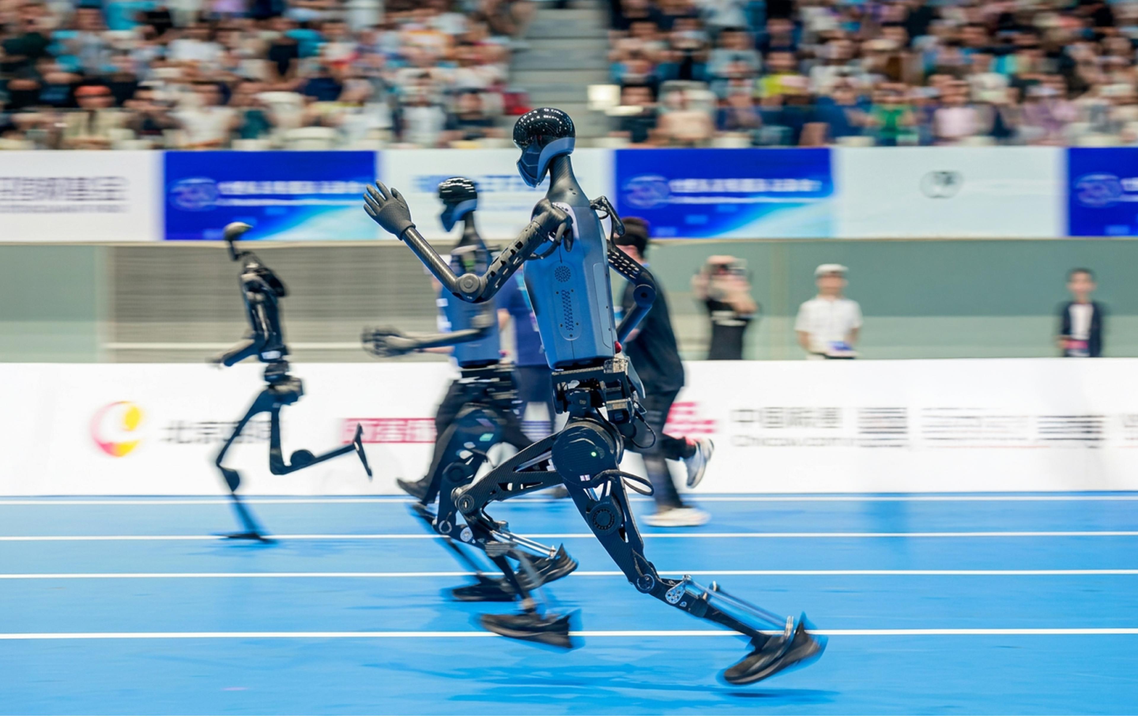 Black humanoid robots sprinting on a blue indoor athletics track in front of a packed stadium crowd