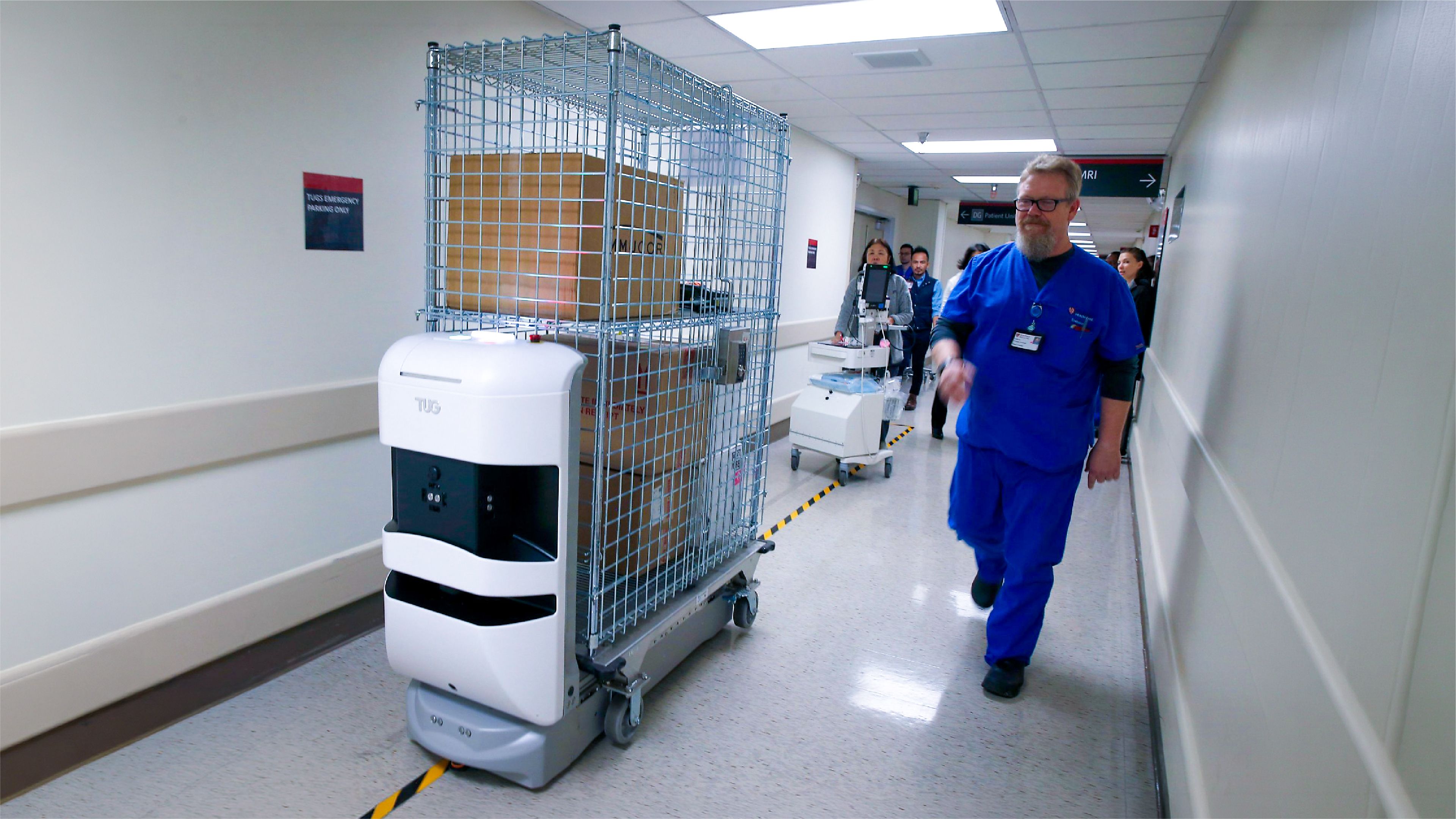 Autonomous TUG robot towing a wire cage cart of supplies along a hospital corridor as a nurse in blue scrubs walks past