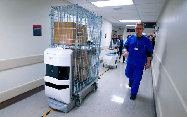 Autonomous TUG robot towing a wire cage cart of supplies along a hospital corridor as a nurse in blue scrubs walks past
