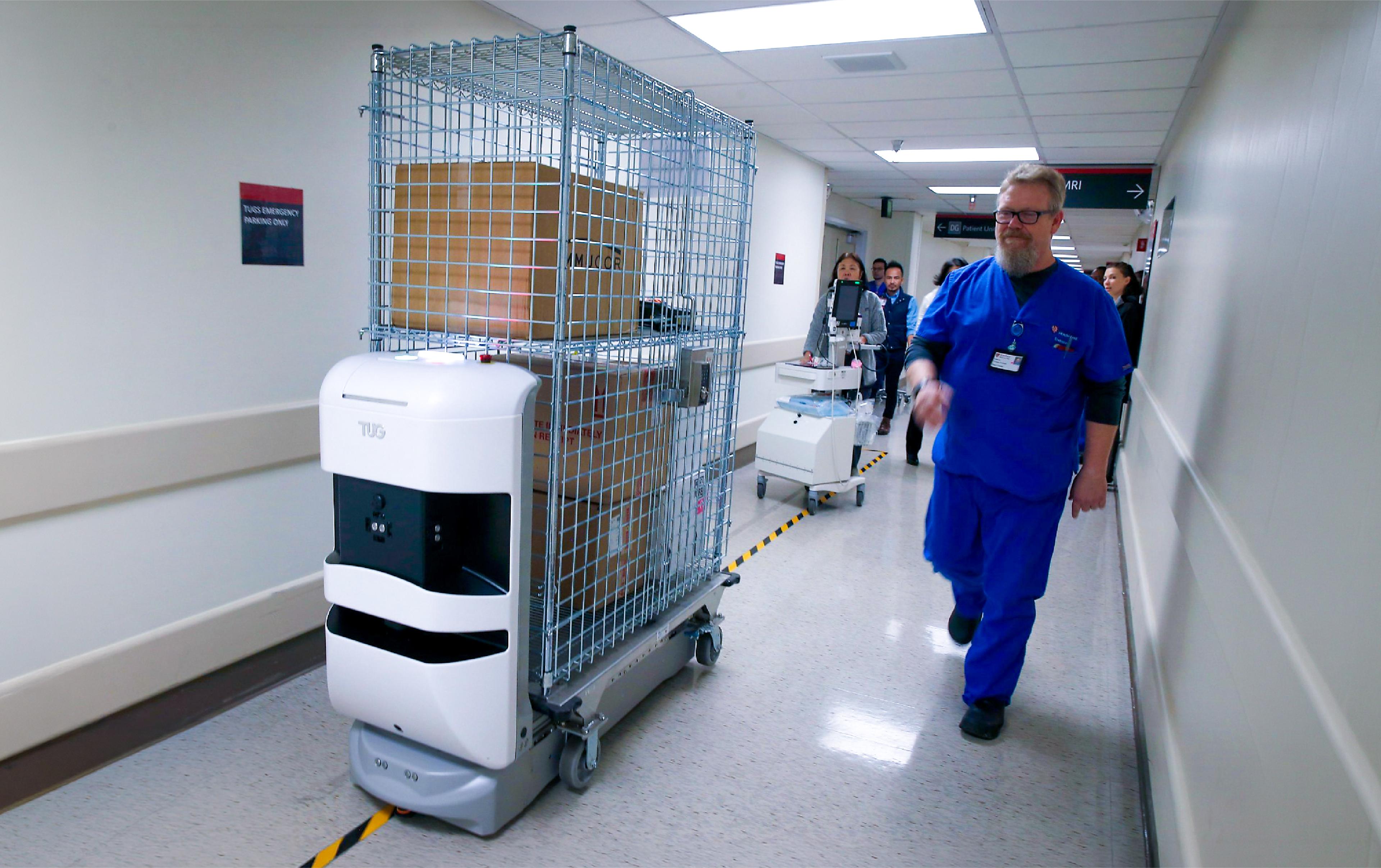 Autonomous TUG robot towing a wire cage cart of supplies along a hospital corridor as a nurse in blue scrubs walks past